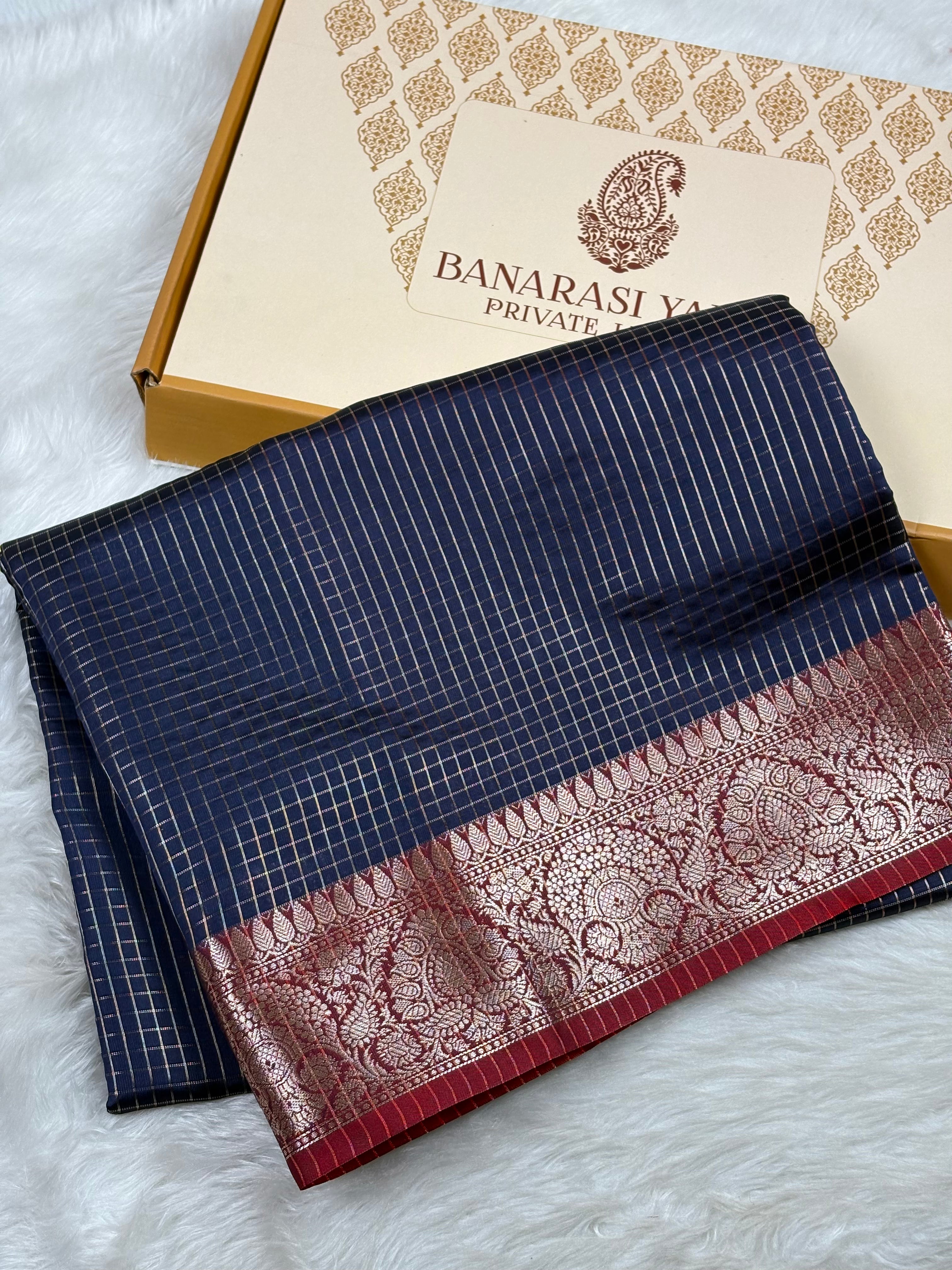 A folded navy blue saree with fine white stripes and a wide red border featuring intricate gold zari weaving, displayed next to its branded Banarasi Yards gift box on a white faux fur surface.