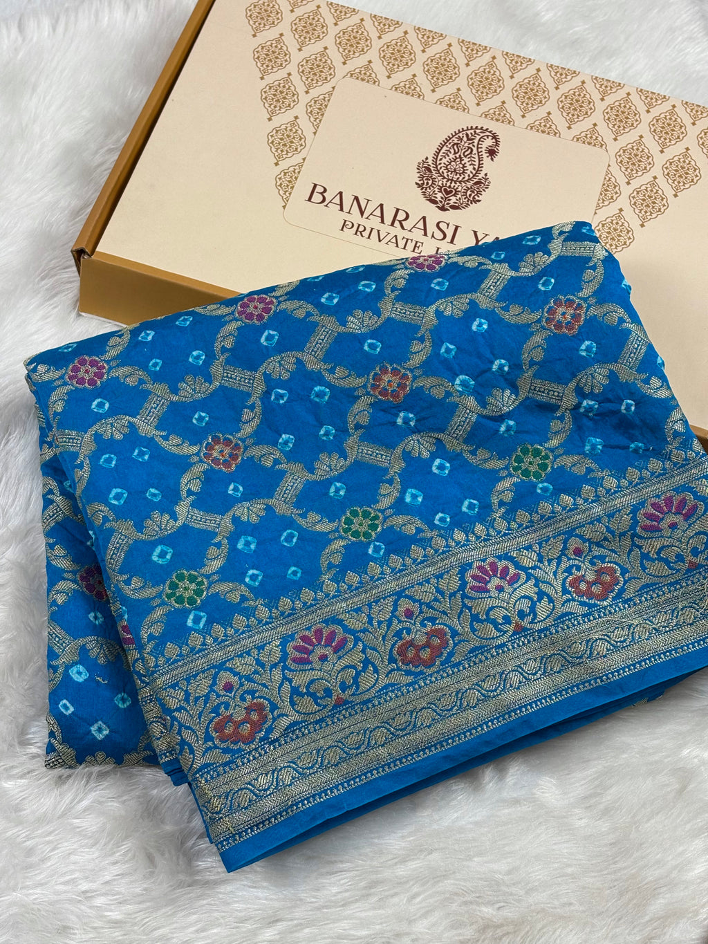 A folded blue Banarasi silk saree with intricate gold zari patterns and floral motifs, displayed next to its packaging box on a white faux fur surface.