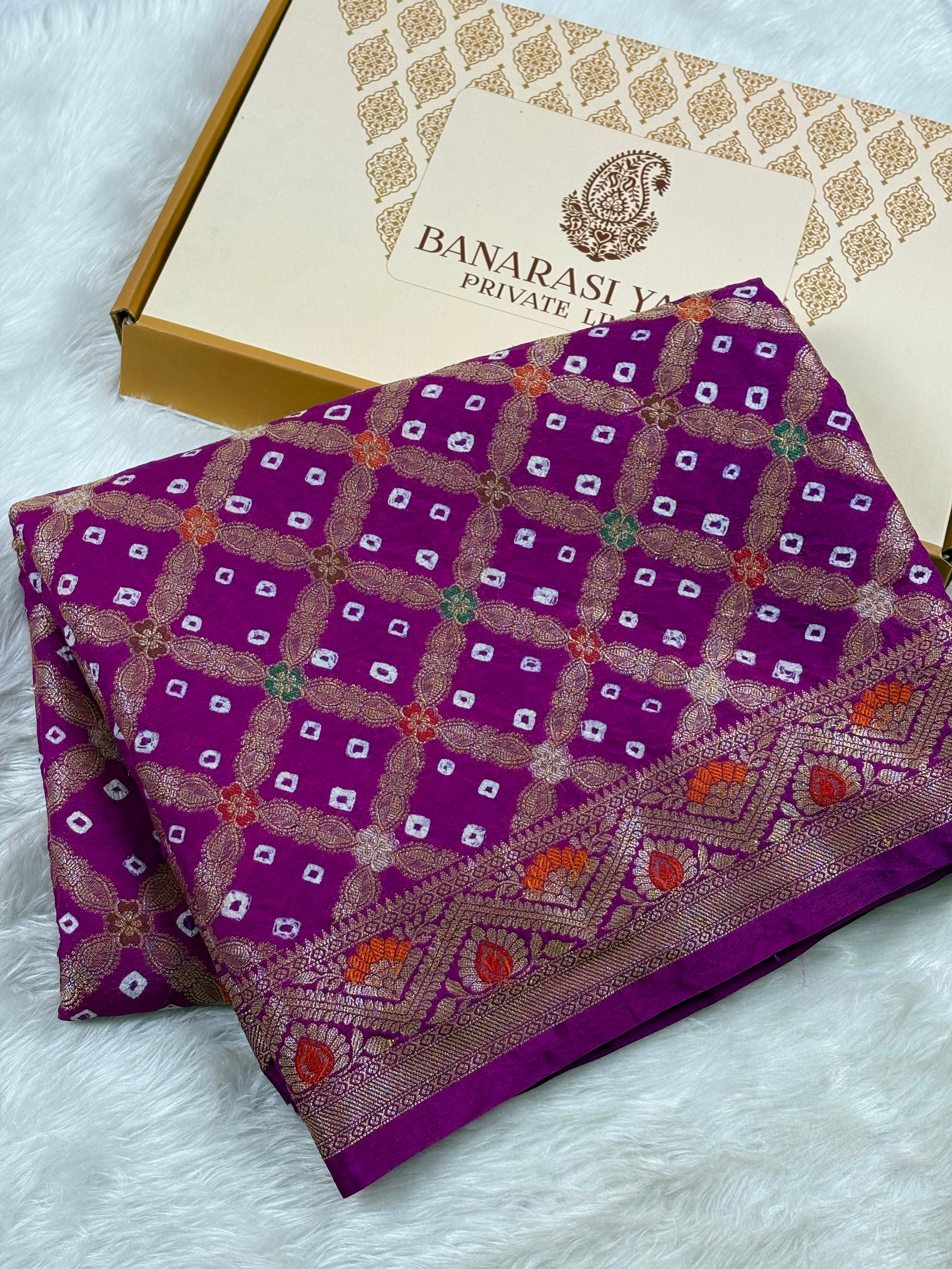 A folded purple Banarasi silk saree with intricate gold, orange, mina weaving and a decorative border, displayed next to its branded gift box on a white faux fur surface.