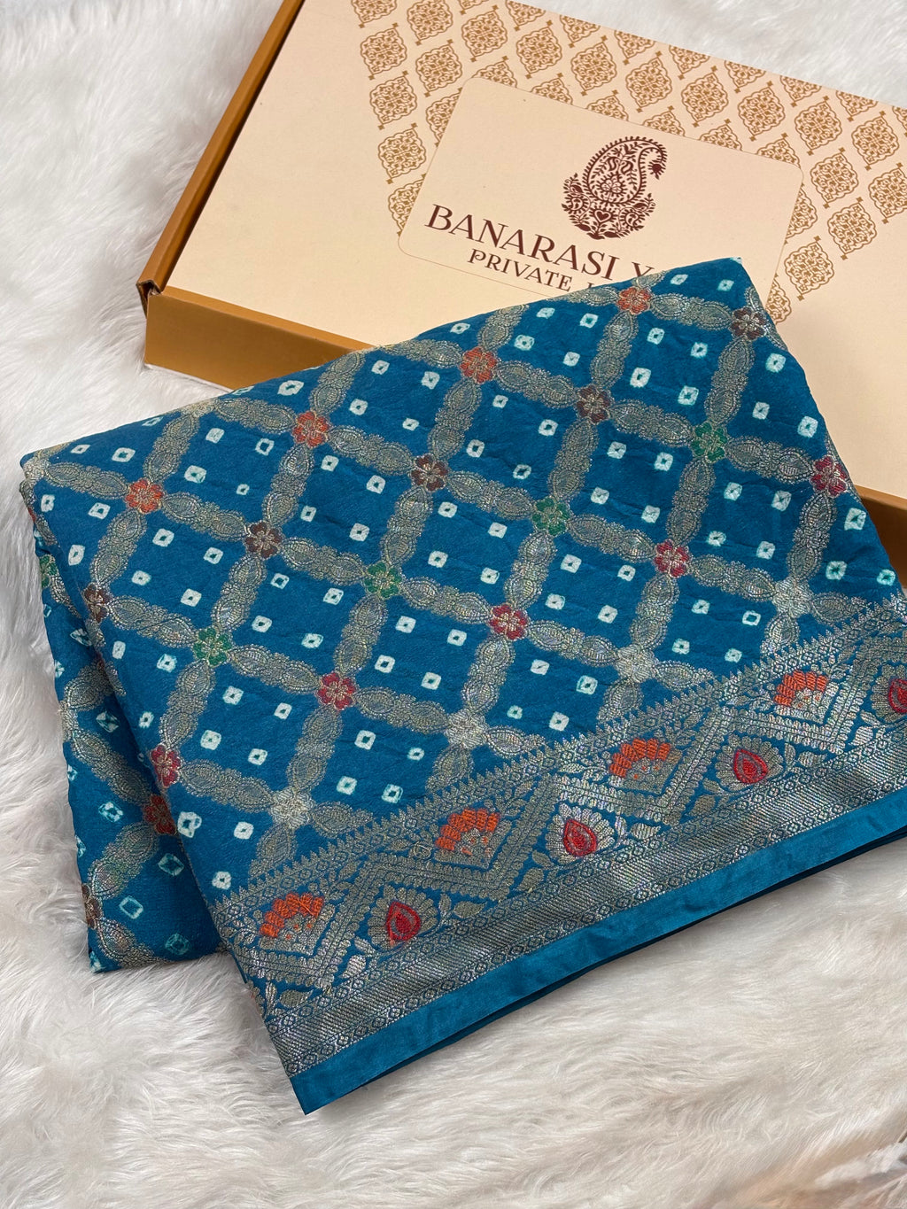 A folded blue patterned saree with metallic geometric and floral motifs, placed on a white faux-fur surface beside a Banarasi Yards box.