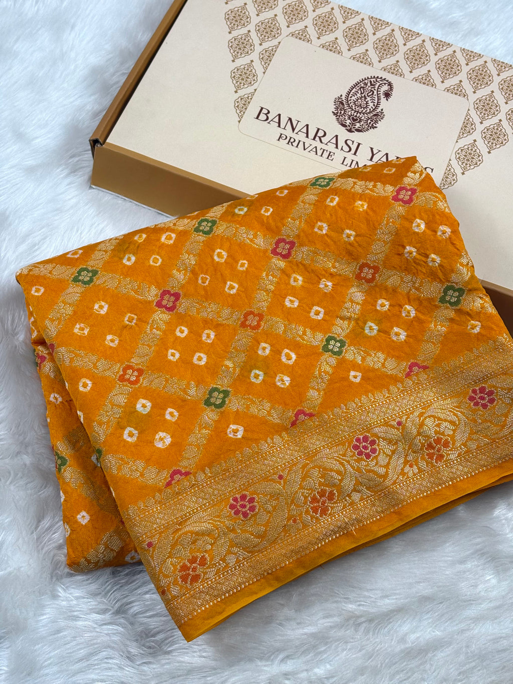 A folded mustard yellow Banarasi saree with intricate gold, red, and green mina woven patterns and a decorative border, displayed next to its branded packaging box on a white faux fur surface.