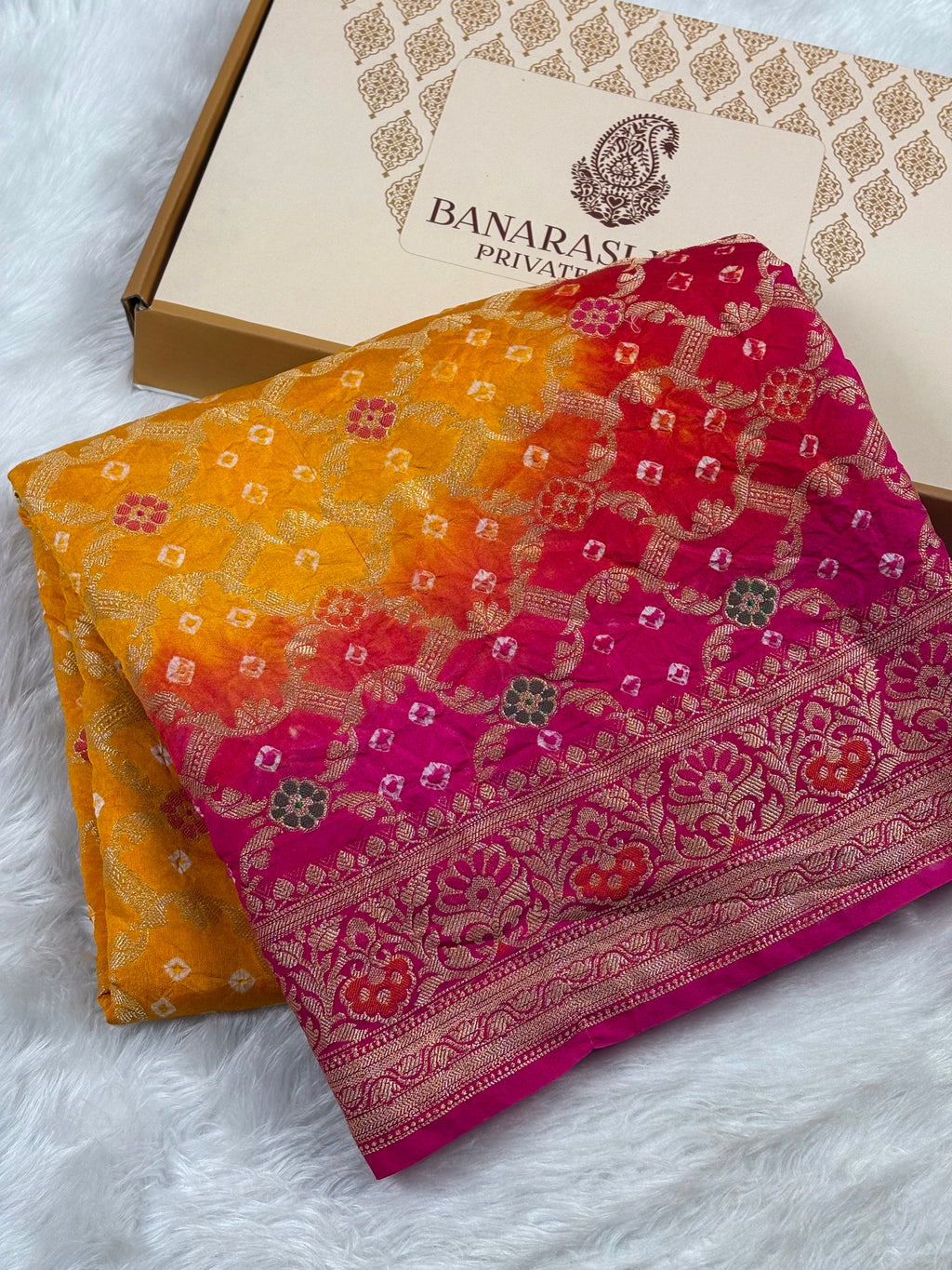 A folded multicolor Banarasi silk saree with a yellow-to-pink gradient and intricate gold zari weaving, displayed next to its branded box on a white faux fur surface.