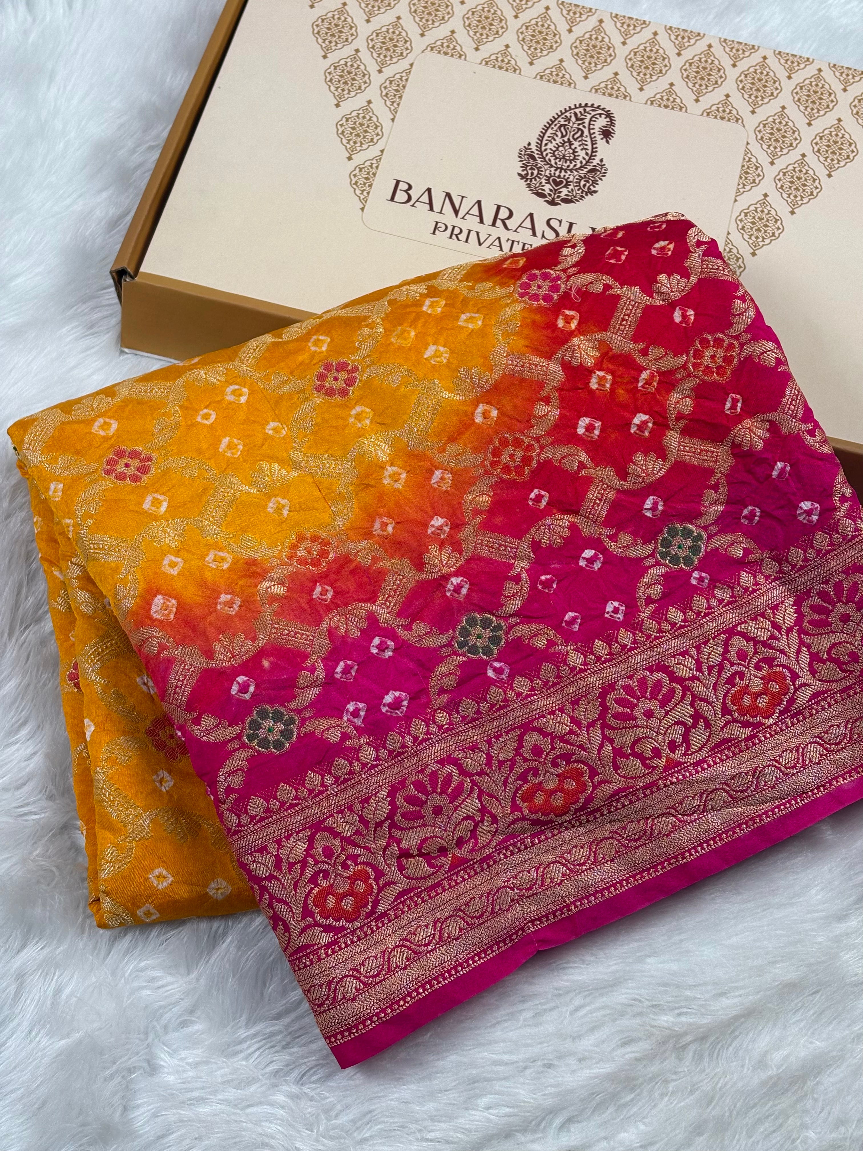 A folded multicolor Banarasi silk saree with a yellow-to-pink gradient and intricate gold zari weaving, displayed next to its branded box on a white faux fur surface.