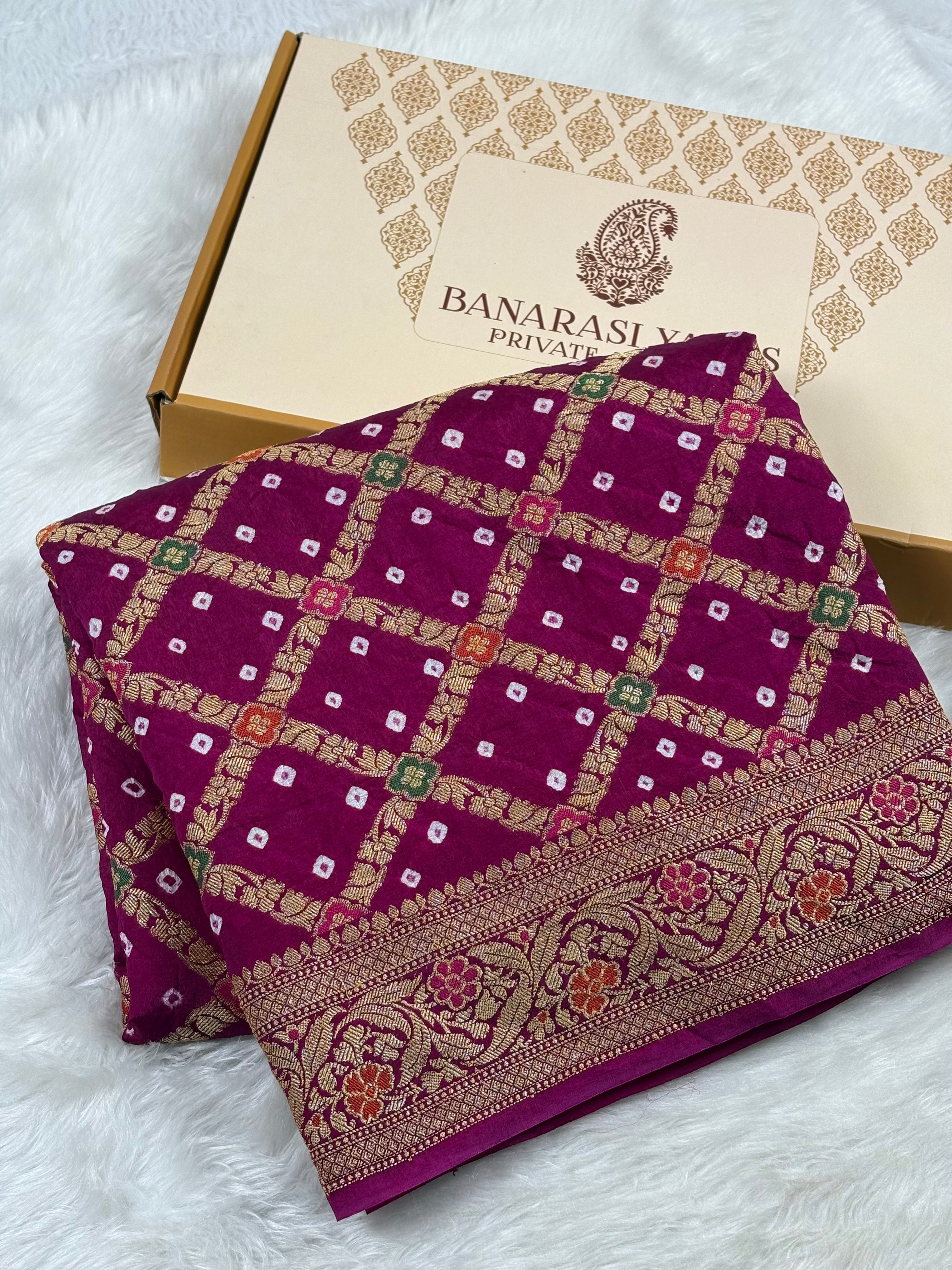 A folded deep magenta purple Banaras silk saree with intricate gold zari weaving and a border, displayed next to its Banarasi Yards brand box on a white faux fur background.