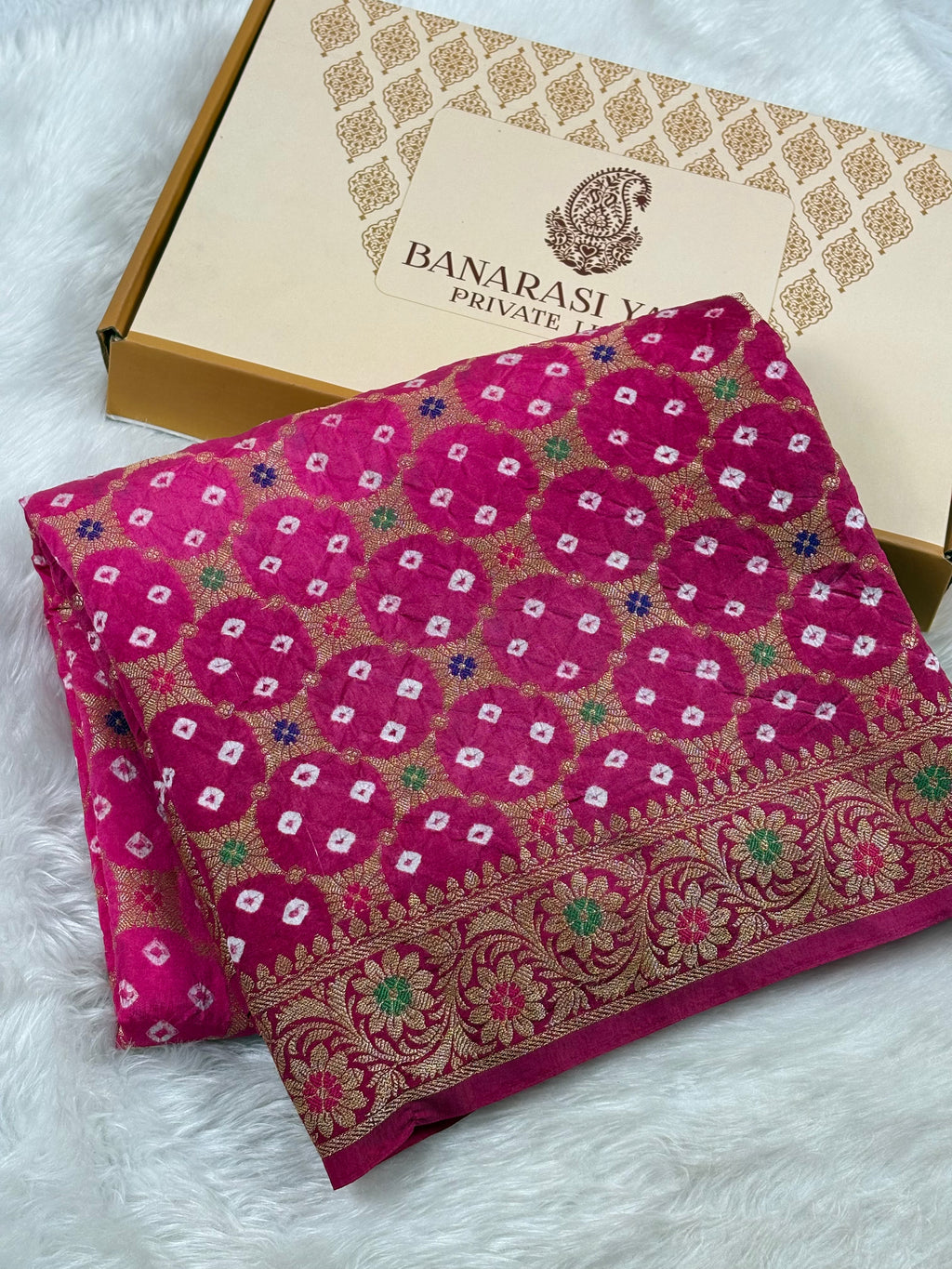 A folded magenta pink Banarasi silk saree with intricate gold, green, and white woven patterns, displayed next to its Banarasi Yards brand box on a white faux fur background.