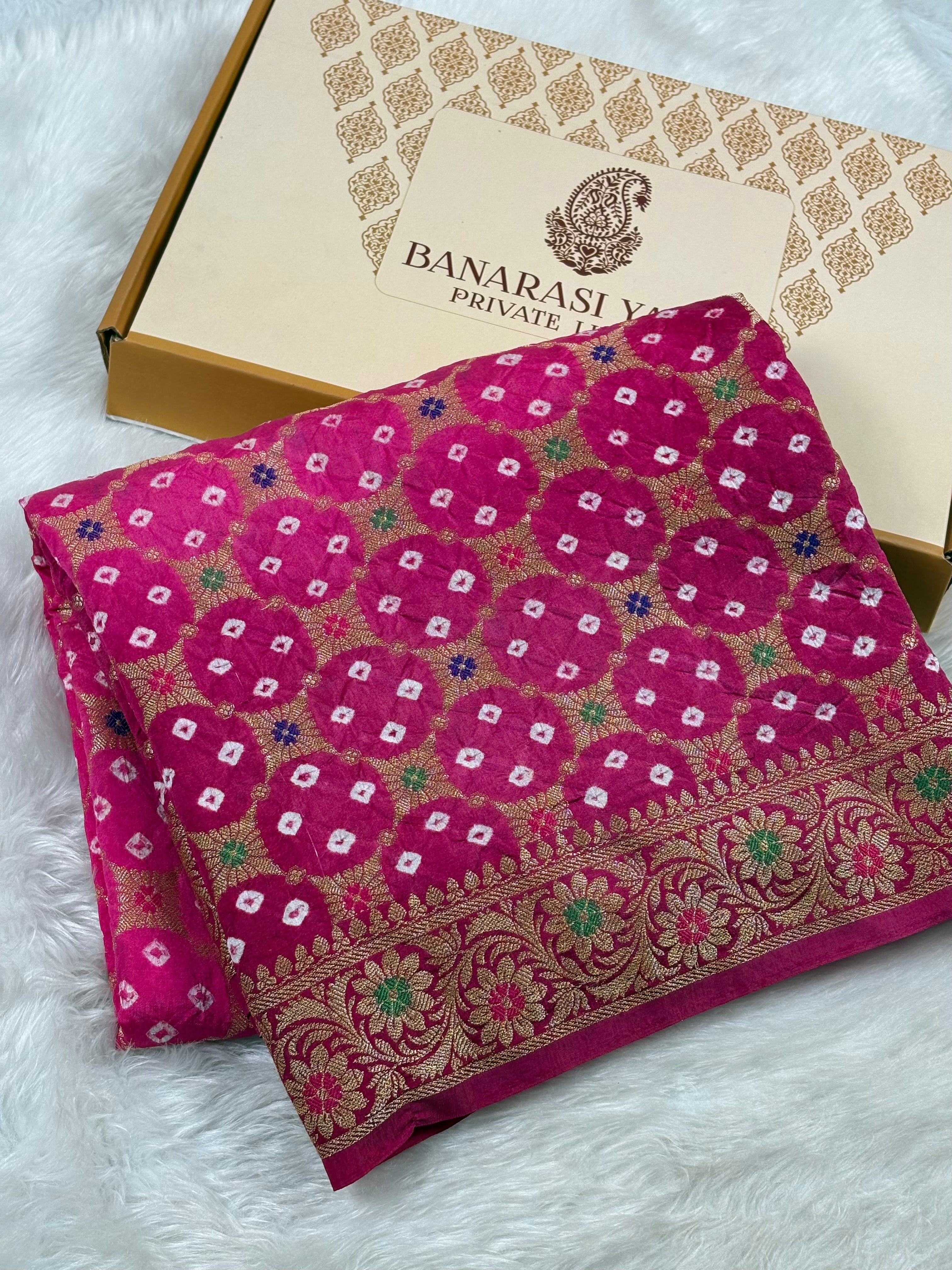 A folded magenta pink Banarasi silk saree with intricate gold, green, and white woven patterns, displayed next to its Banarasi Yards brand box on a white faux fur background.