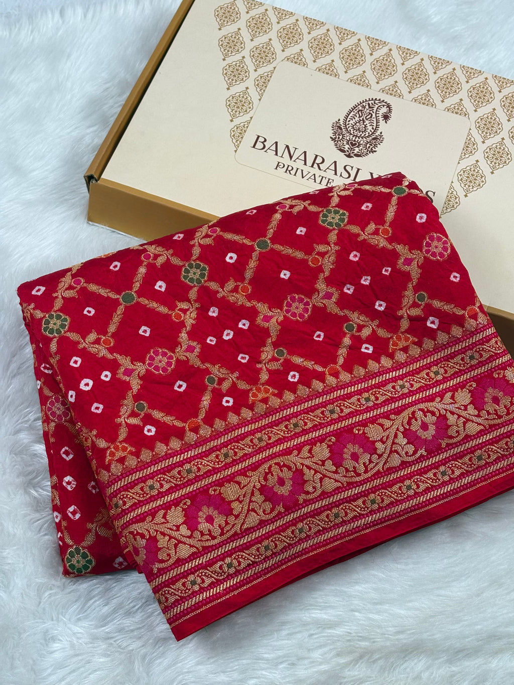 A folded red Banarasi silk saree with intricate gold, pink, and green woven patterns, displayed next to its packaging box on a white faux fur surface.