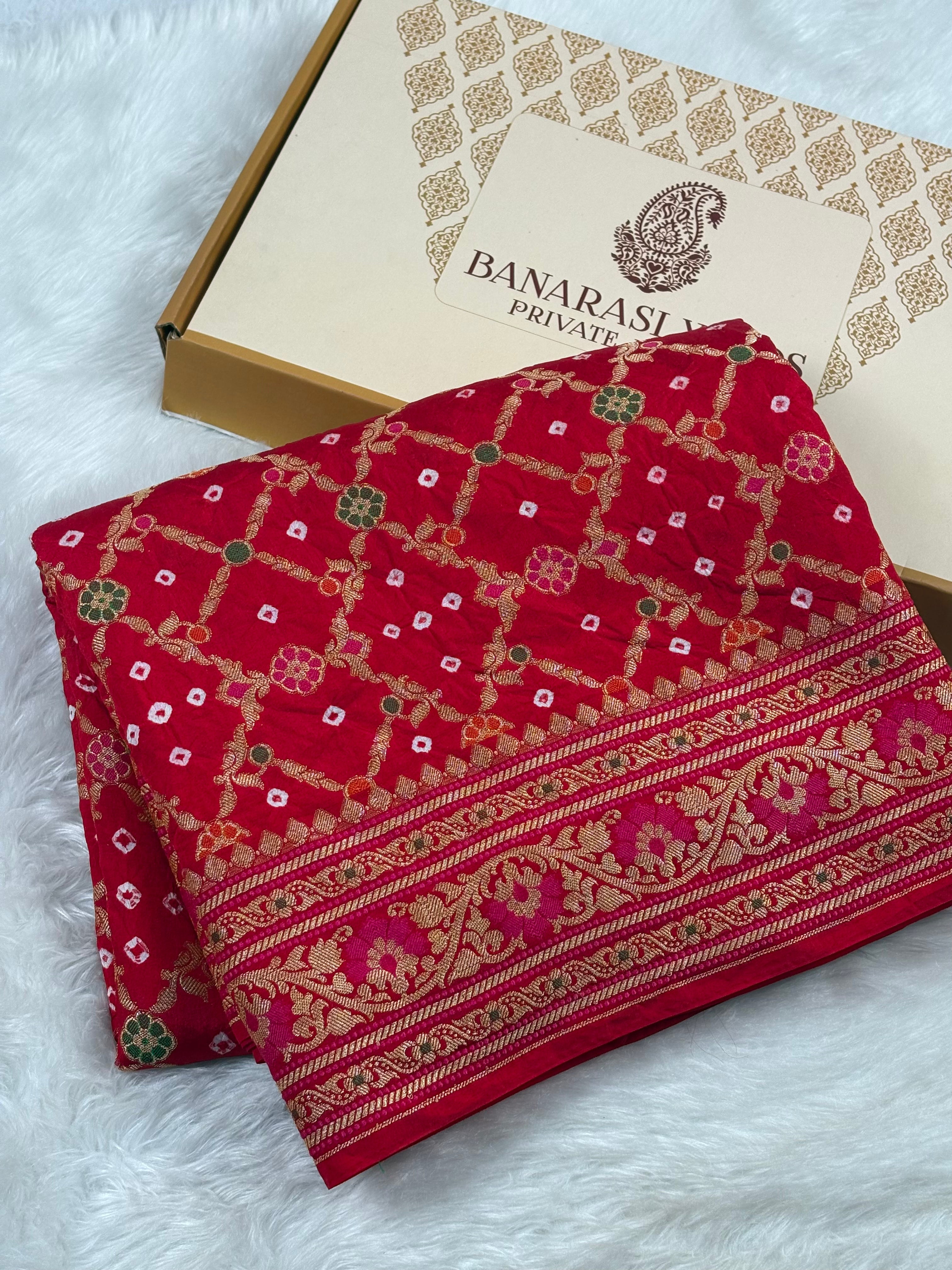 A folded red Banarasi silk saree with intricate gold, pink, and green woven patterns, displayed next to its packaging box on a white faux fur surface.