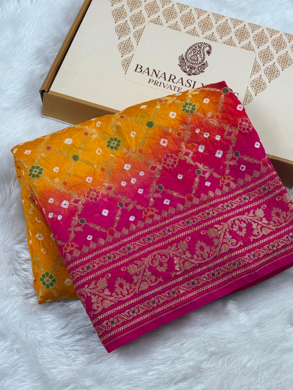 A folded Banarasi silk saree with a vibrant orange-to-pink gradient and intricate gold zari weaving, displayed next to its branded box on a white faux fur surface.