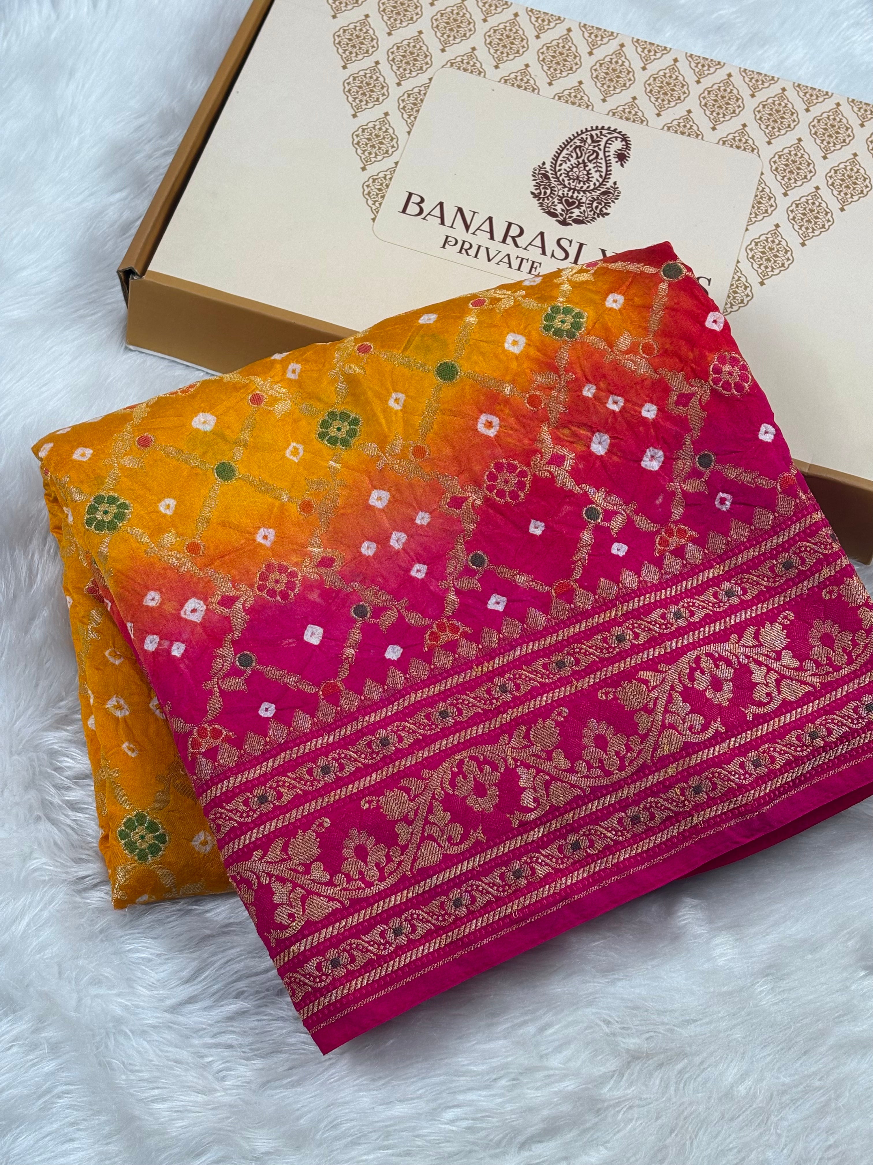 A folded Banarasi silk saree with a vibrant orange-to-pink gradient and intricate gold zari weaving, displayed next to its branded box on a white faux fur surface.