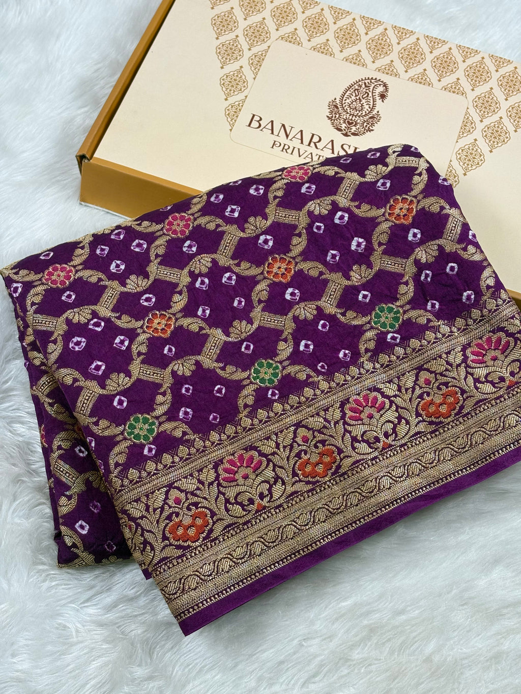 A folded purple Banarasi silk saree with intricate gold zari weaving and colorful floral motifs, displayed next to its branded packaging box on a white faux fur surface.