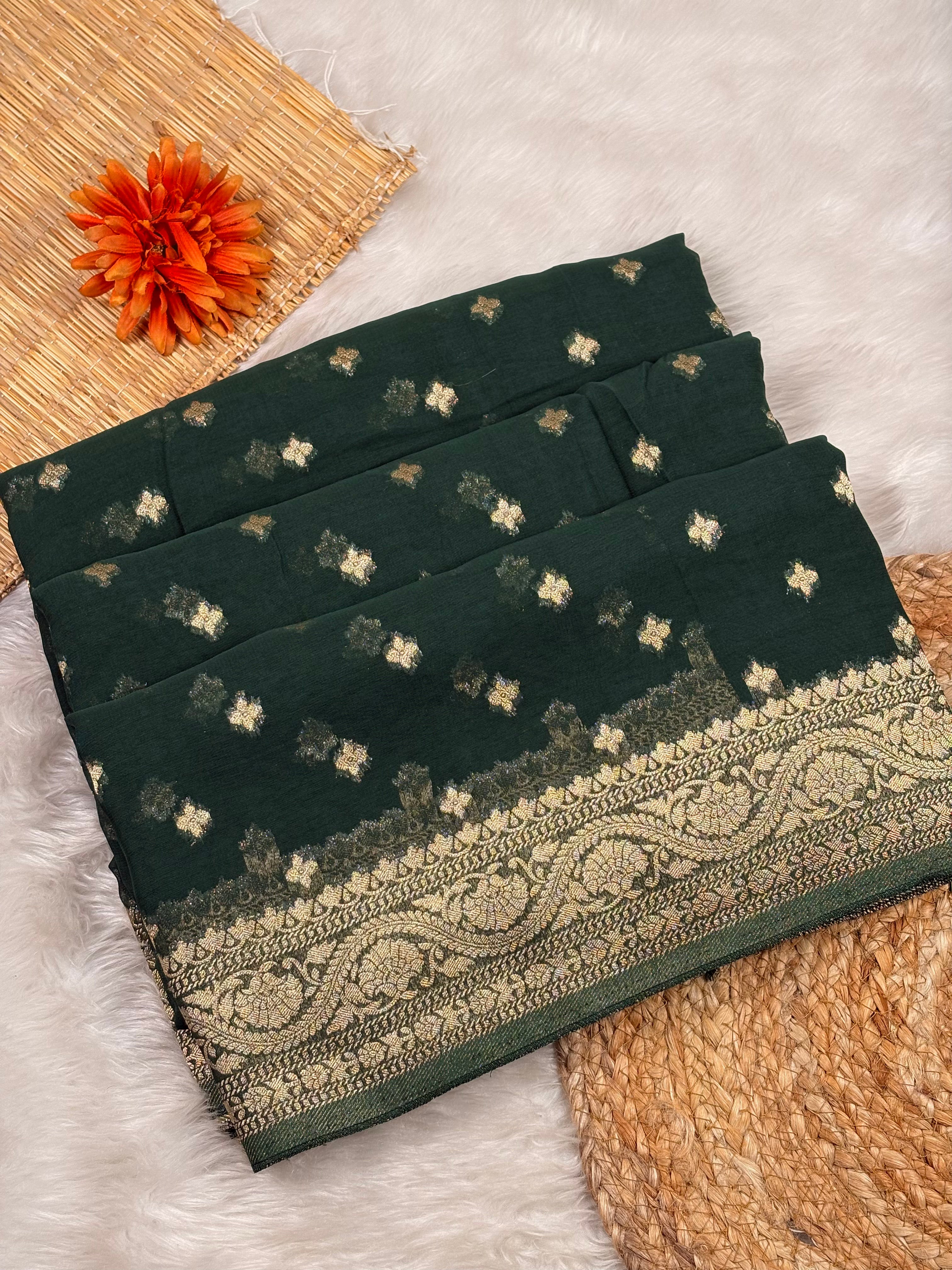 A folded dark forest green saree, decorated with small beige and white floral motifs and a wide beige paisley border. The saree is styled on a white faux fur surface with a woven mat, a decorative orange flower, and a jute mat as props.