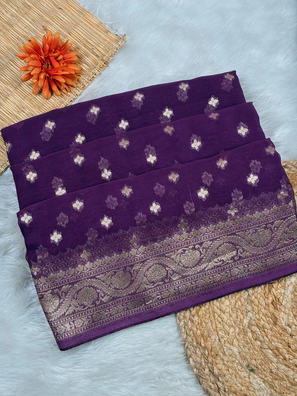 A folded purple saree with small metallic floral motifs and a wide ornate border, styled on a light faux-fur surface with woven props and an orange flower.