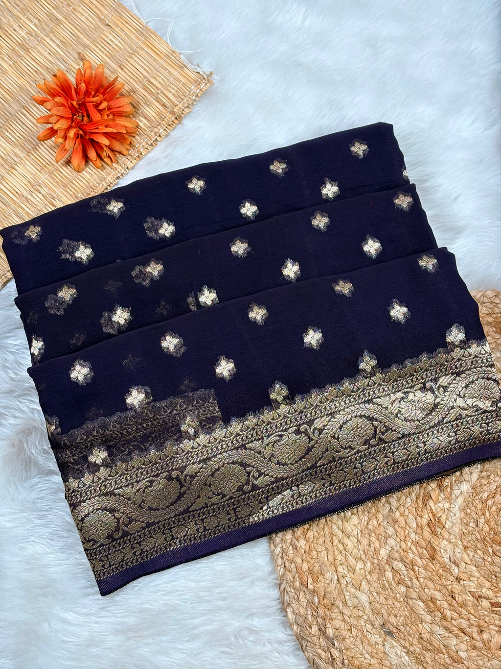 A folded dark navy blue saree with scattered gold floral motifs and a wide ornate gold border, styled on a white faux-fur surface with woven props and an orange flower.