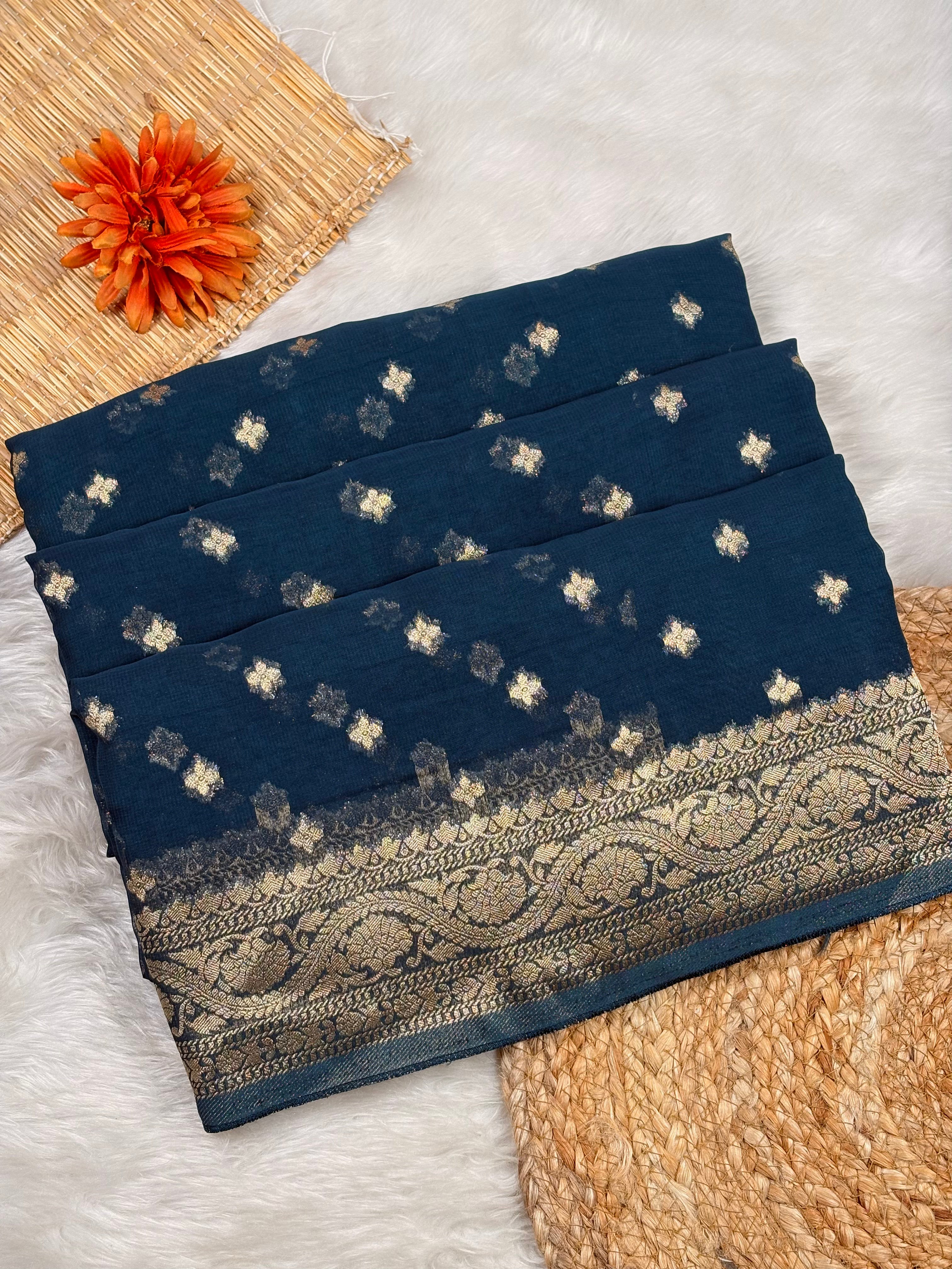 A folded navy blue saree, decorated with small metallic gold floral motifs and a wide ornate gold border, styled on a white faux fur surface with a woven mat and orange flower prop.
