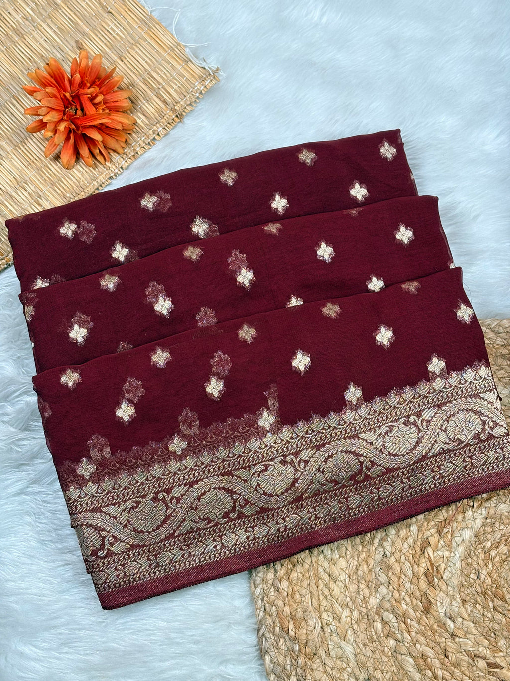 A folded deep maroon with metallic gold floral motifs and a wide ornate gold border, styled on a white faux-fur surface with woven mats and an orange flower prop.