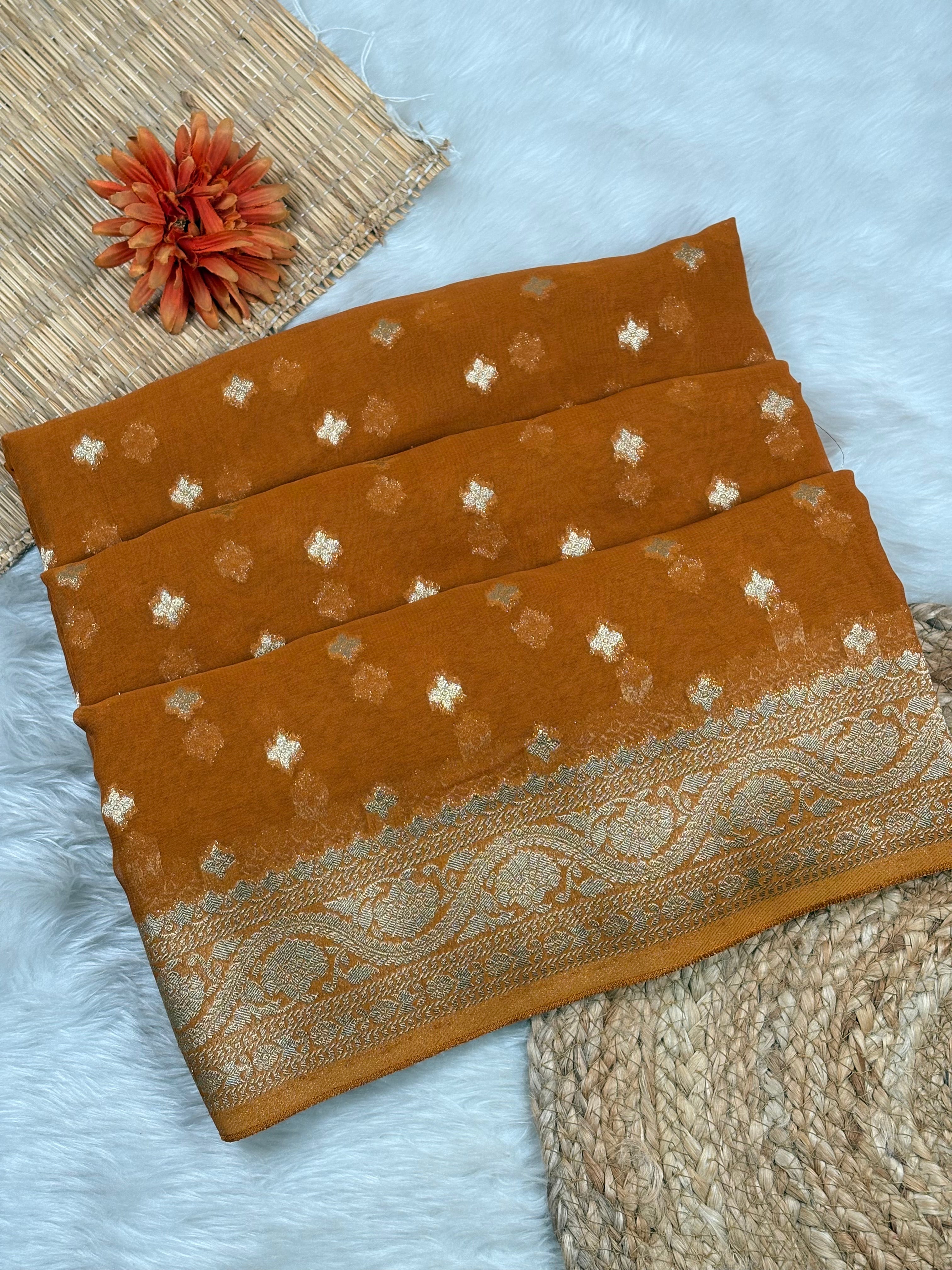 A folded ochre-brown with small white floral motifs and a wide ornate beige paisley border, styled on a white faux-fur surface with woven props and a dried orange flower.
