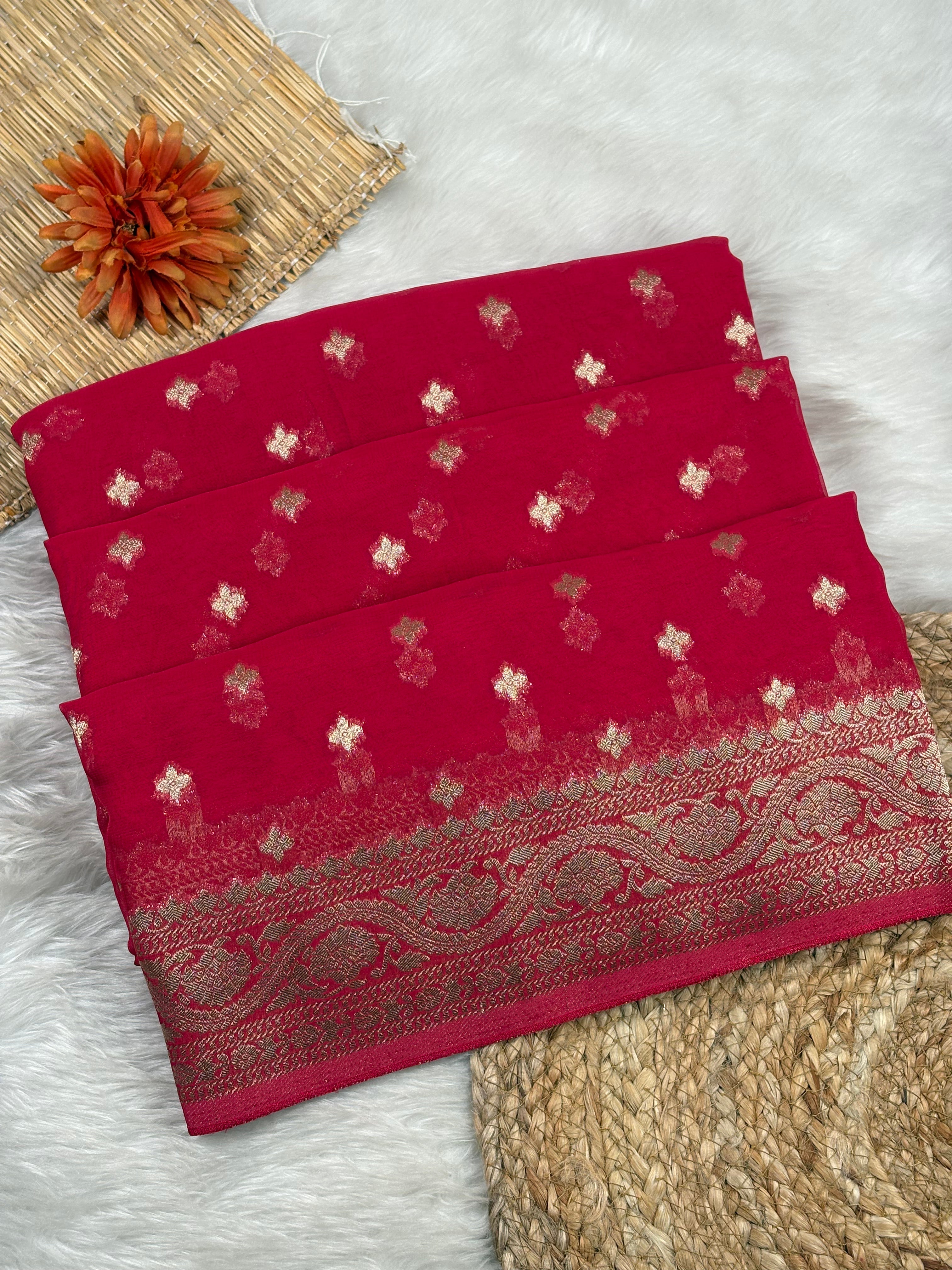 A folded red saree with small metallic gold floral motifs and a wide ornate gold border, displayed on a white faux-fur surface with woven props.
