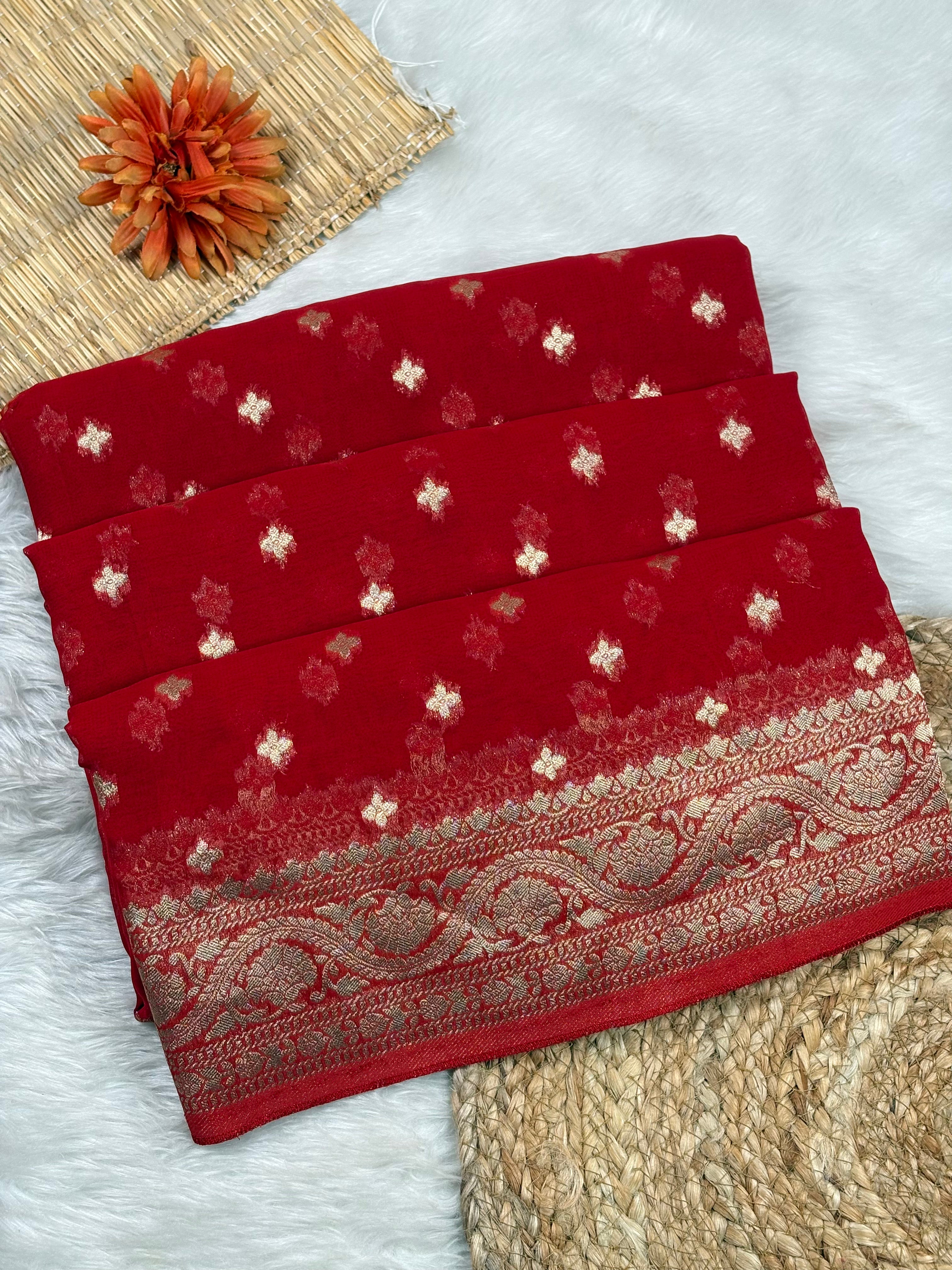 A folded red saree piece with small metallic floral motifs and a wide ornate brocade border, arranged on a white faux-fur surface with woven mat props.