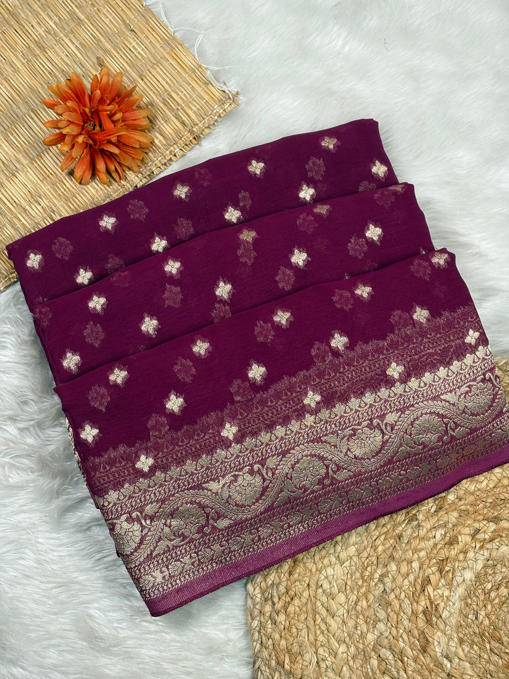 A folded deep purple saree with small metallic floral motifs and a wide ornate gold border, styled on a white faux-fur surface with woven mat props and an orange flower.
