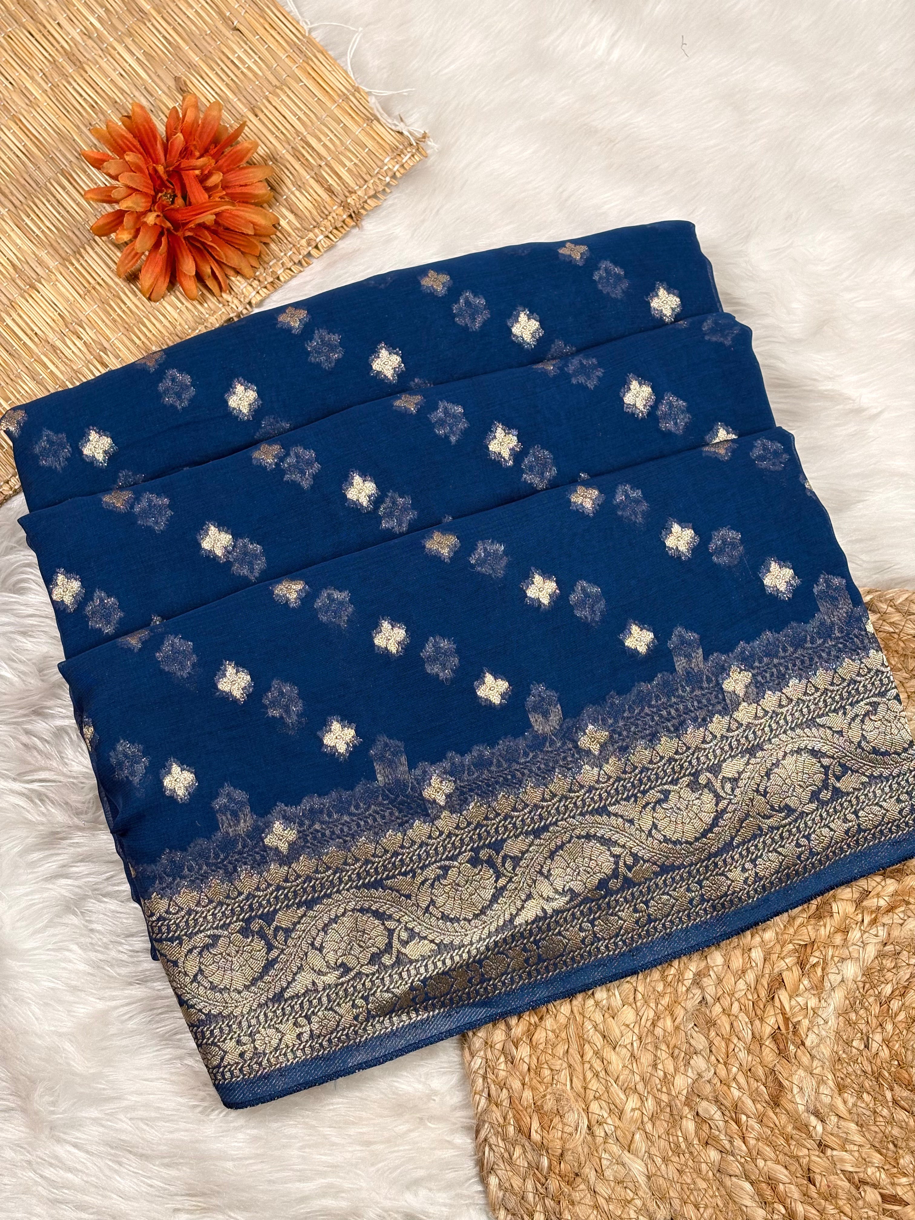 A folded navy blue saree with small white floral motifs and a wide ornate gold border, styled on a white faux-fur surface with woven mats and an orange flower prop.