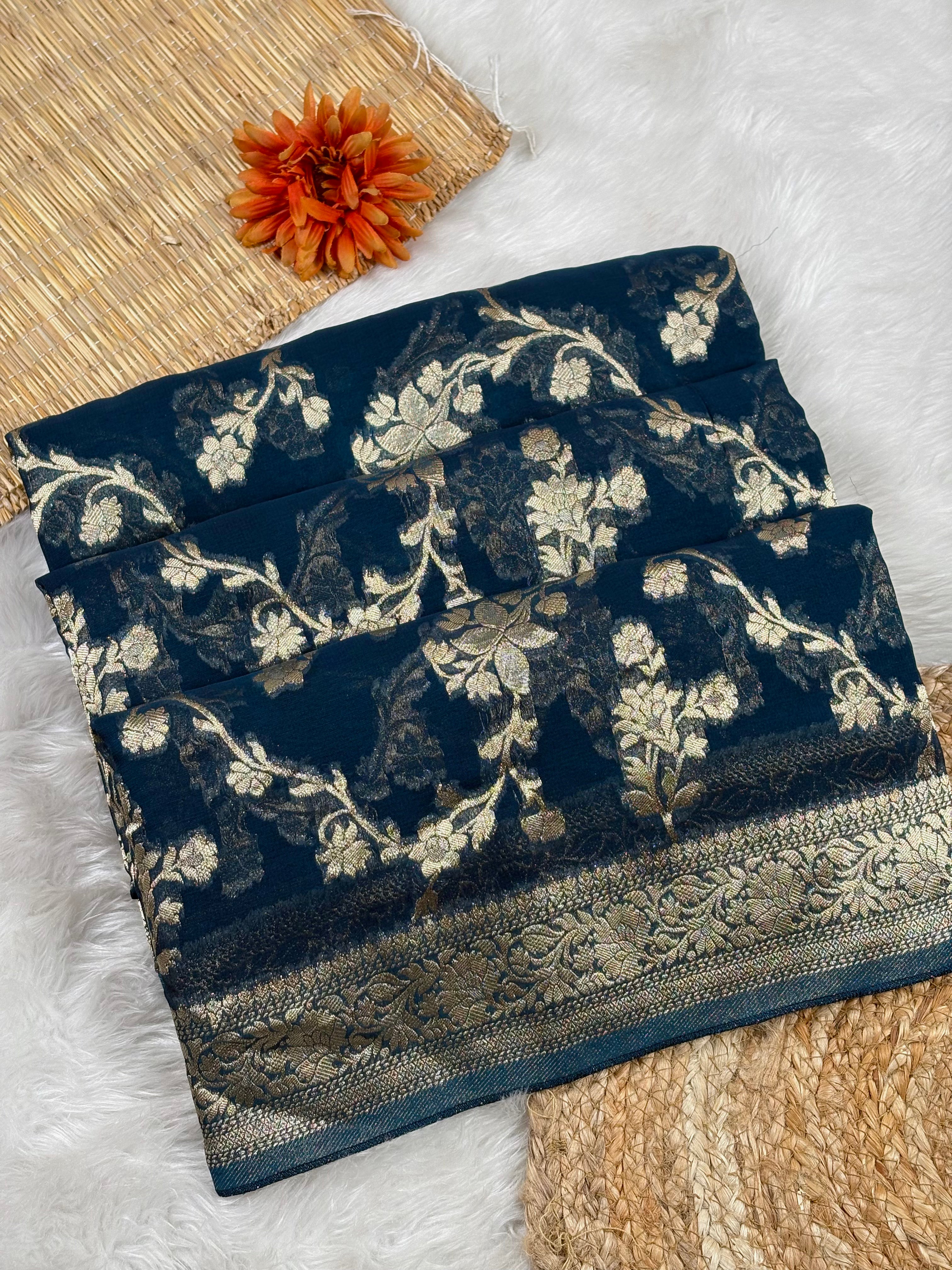 A folded dark navy blue saree with intricate gold floral brocade patterns and a decorative border, styled on a white faux-fur surface with a woven mat and orange flower prop.