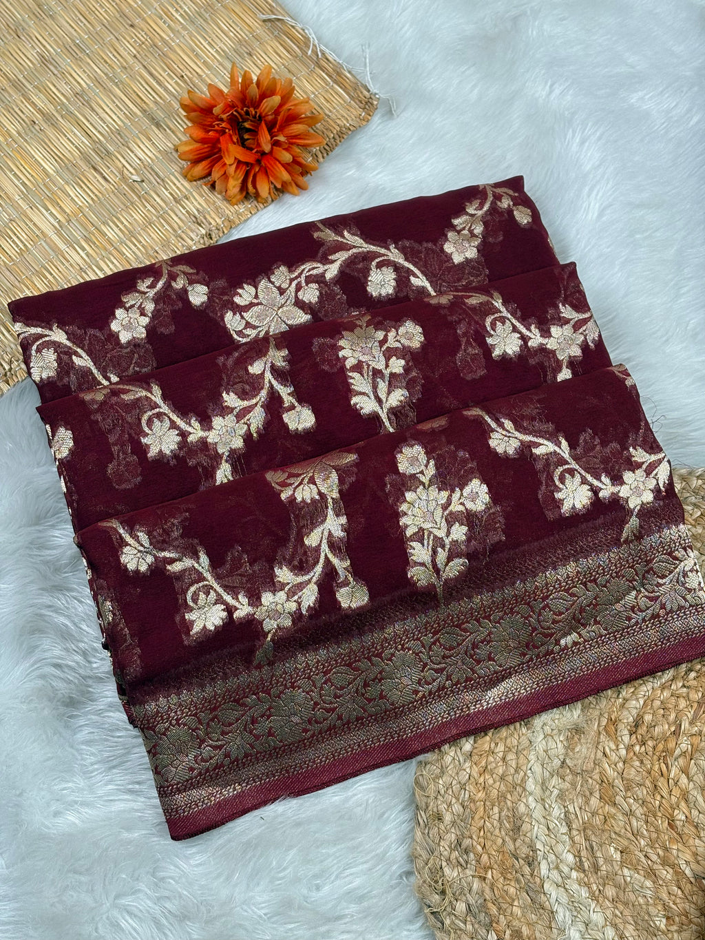 A folded deep maroon saree with gold floral weaving and a patterned border, styled on a white faux-fur surface with a woven mat and an orange flower prop.