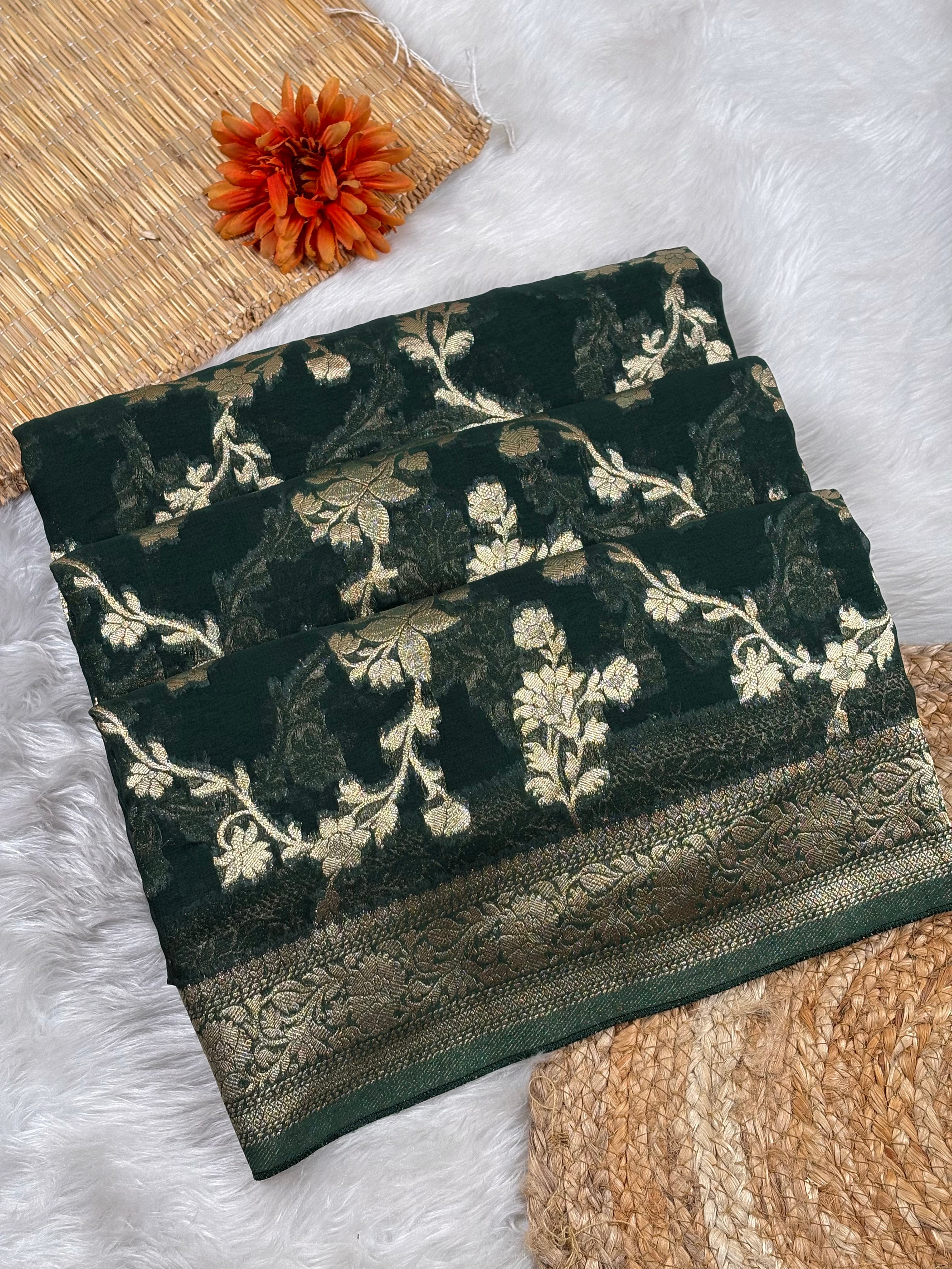 A folded dark green saree with intricate gold floral weaving and a wide metallic gold border, styled on a white faux fur surface with a woven mat and an orange flower.