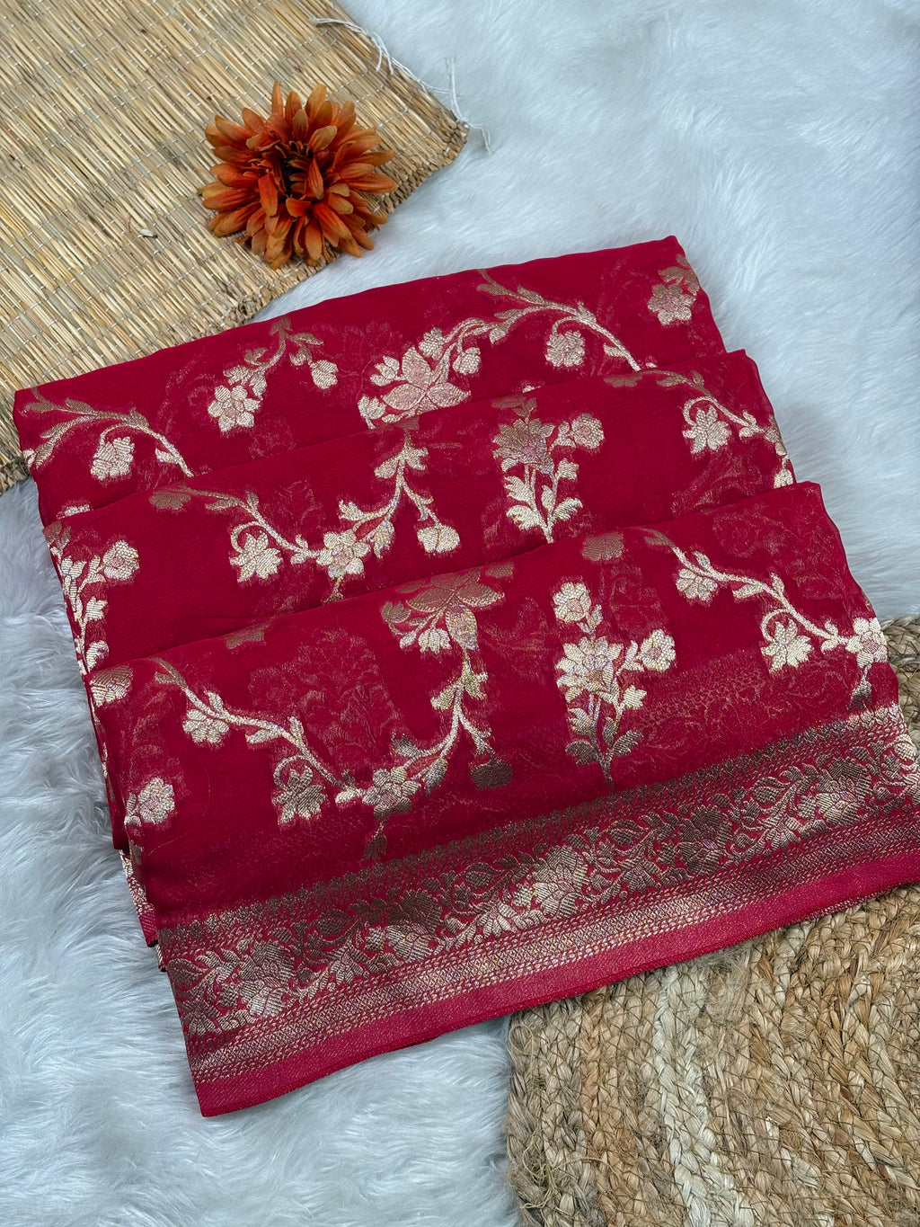 A folded deep red saree, featuring intricate gold floral and vine patterns woven across the surface and a decorative gold border. The fabric is styled in a flat-lay arrangement on a white faux fur surface, alongside a woven mat, a dried orange flower, and a textured beige rug.