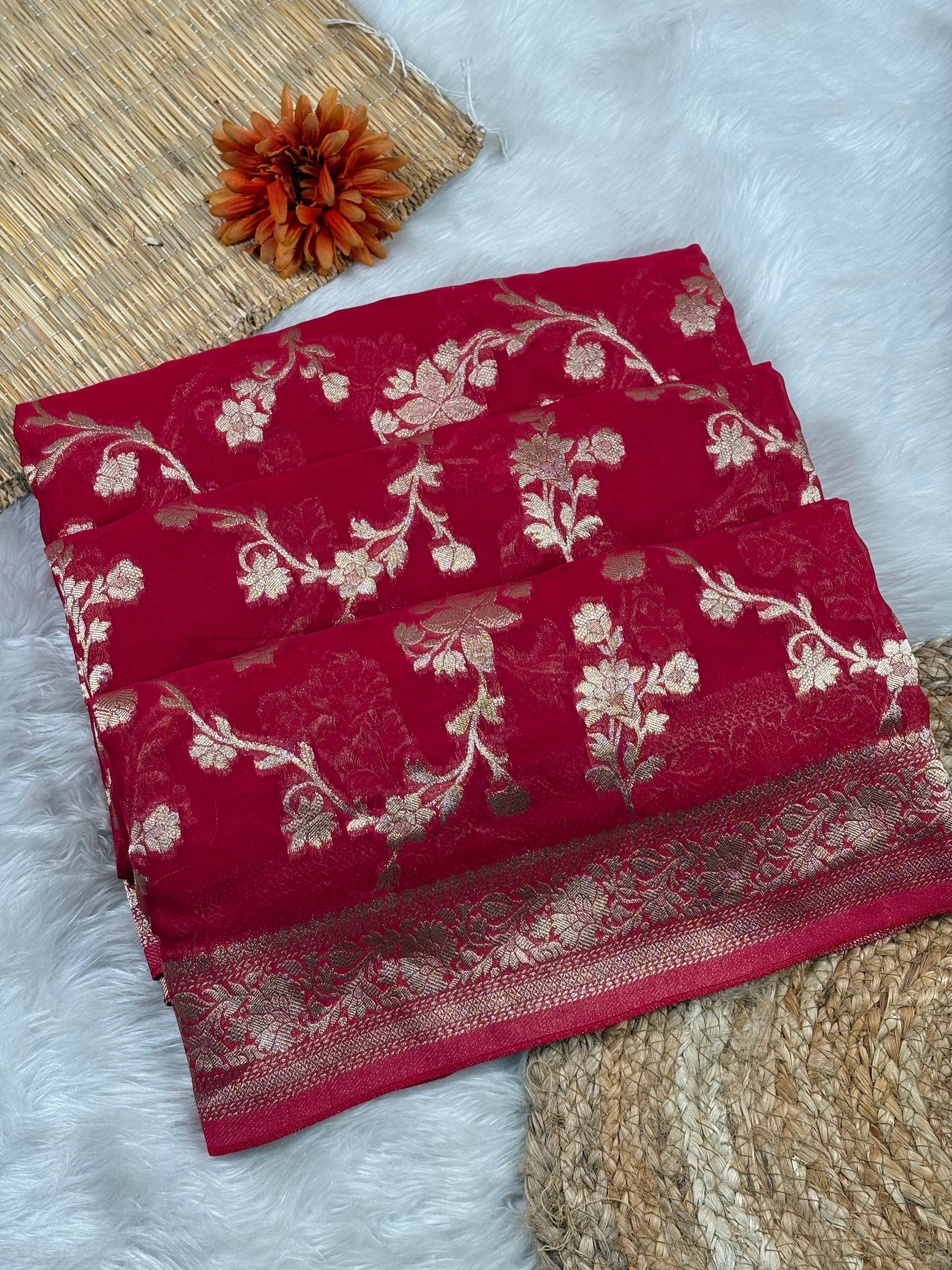 A folded deep red saree, featuring intricate gold floral and vine patterns woven across the surface and a decorative gold border. The fabric is styled in a flat-lay arrangement on a white faux fur surface, alongside a woven mat, a dried orange flower, and a textured beige rug.