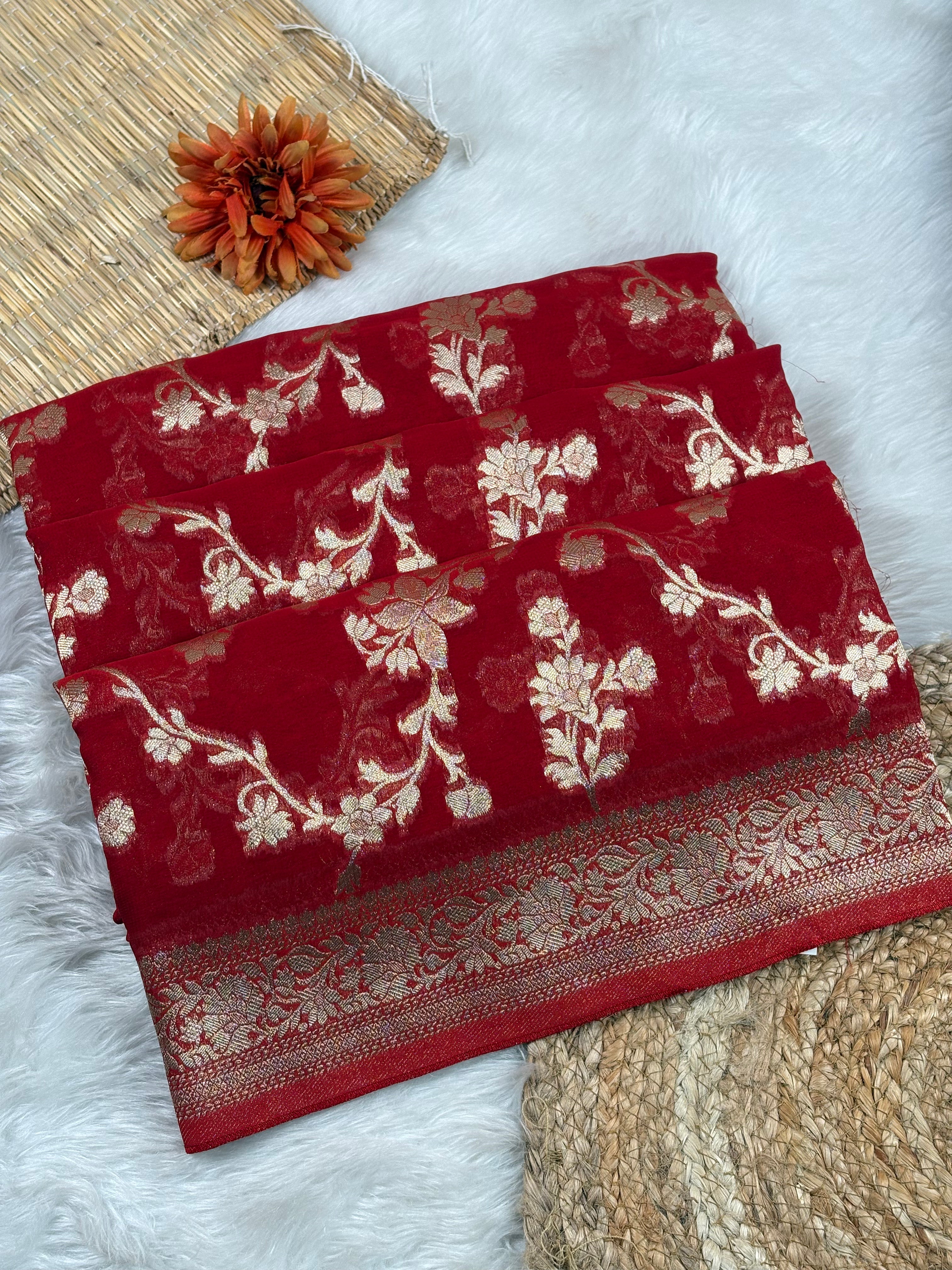 A folded deep red saree with intricate gold floral and vine patterns, displayed on a white faux fur surface with decorative woven mats and a small orange flower.