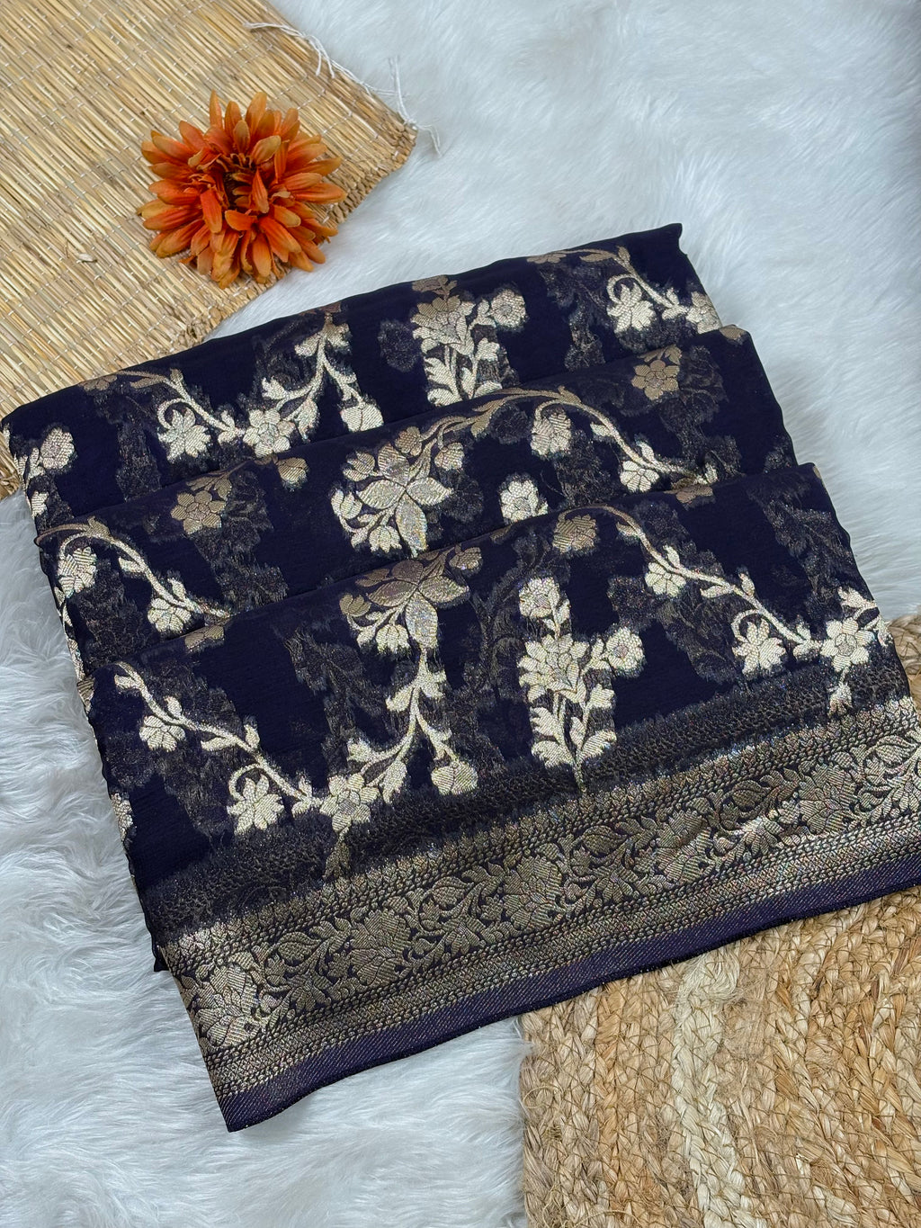 A folded dark navy blue saree with intricate gold floral weaving and a decorative border, styled on a white faux-fur surface with a woven mat, a dried orange flower, and a textured beige textile.
