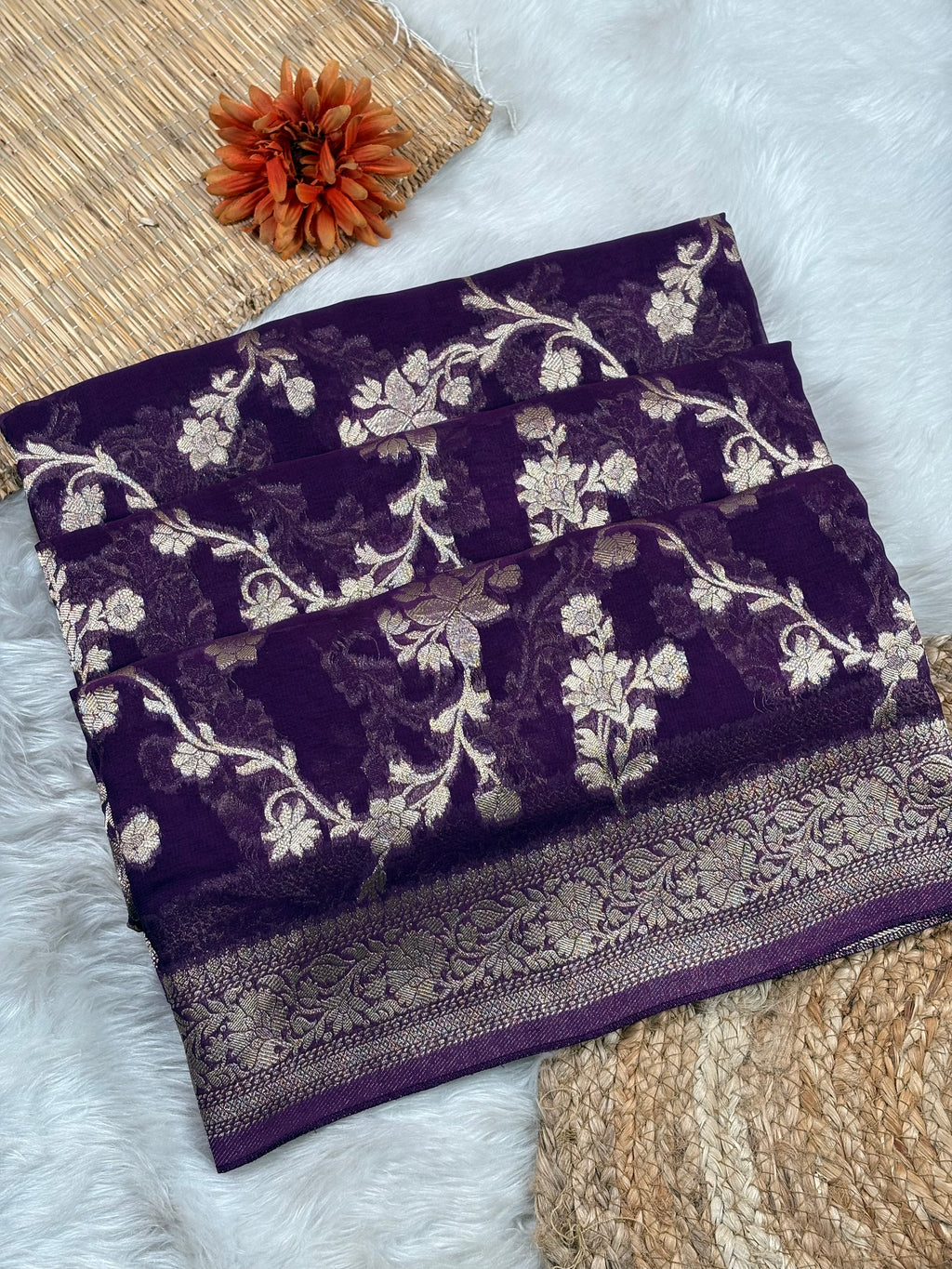 A folded dark purple saree with intricate sgold floral weaving and a decorative border, styled on a white faux-fur surface with a woven mat and an orange flower prop.