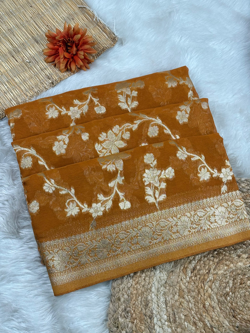 A folded mustard yellow saree with cream floral weaving and a decorative border, styled on a white faux-fur surface with a woven mat and dried flower prop.