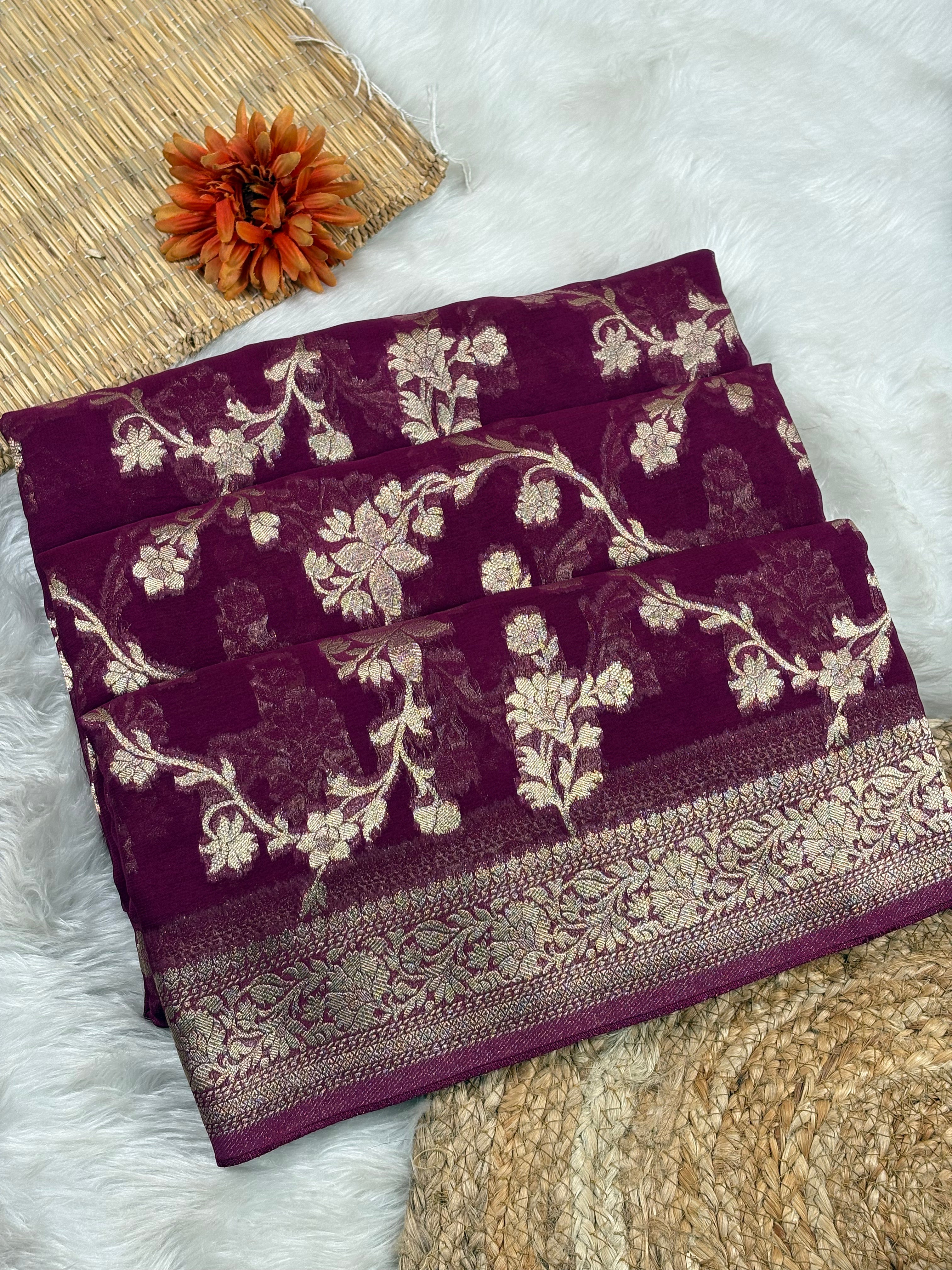 A folded deep purple saree with intricate gold floral weaving and a decorative border, styled on a white faux-fur surface with woven mats and an orange flower accent.