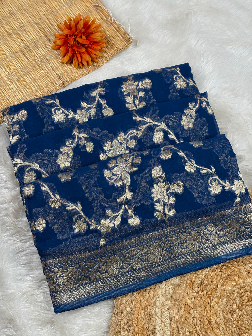 A folded navy blue saree with intricate gold floral weaving and a decorative metallic border, styled on a white faux-fur surface with woven mats and an orange flower accent.