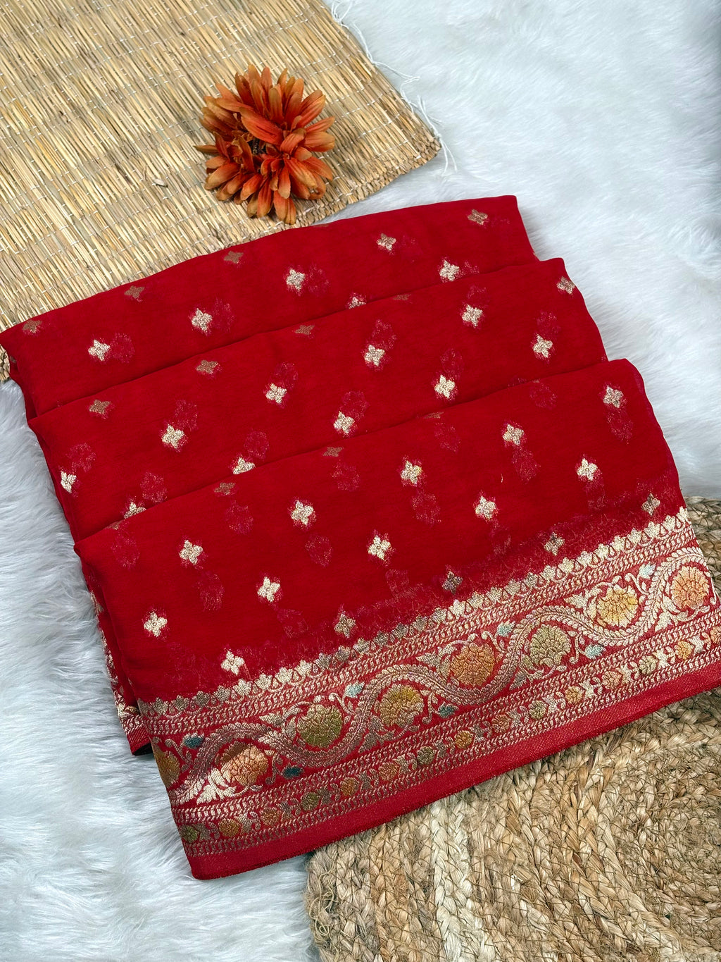 A folded red saree, featuring small metallic gold floral motifs and an ornate gold border, displayed on a white faux fur surface with a woven mat and dried orange flower as props.