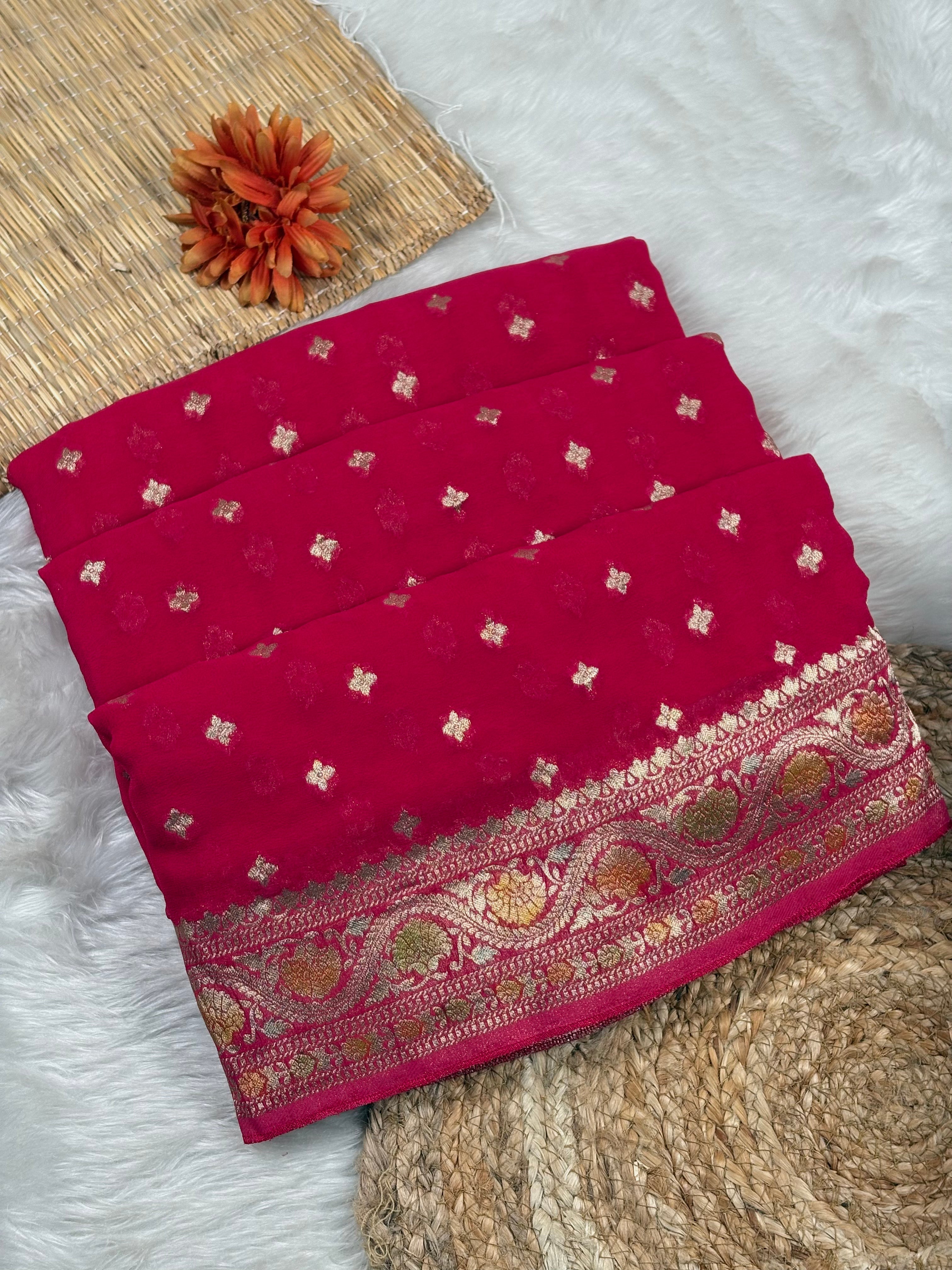 A folded deep magenta pink saree with small gold floral motifs and a wide ornate gold border with handbrush mina, displayed on a white faux fur surface with a woven mat and dried orange flower as props.