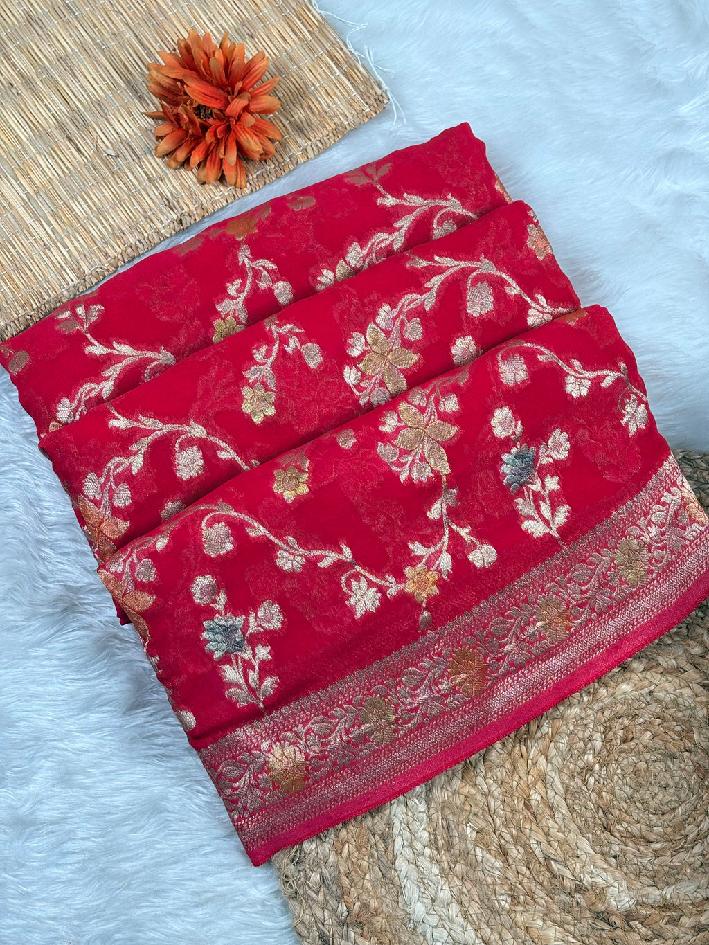 A folded red saree with ornate floral brocade and metallic gold and silver patterns, displayed on a white faux fur surface with a woven mat and a decorative orange flower.