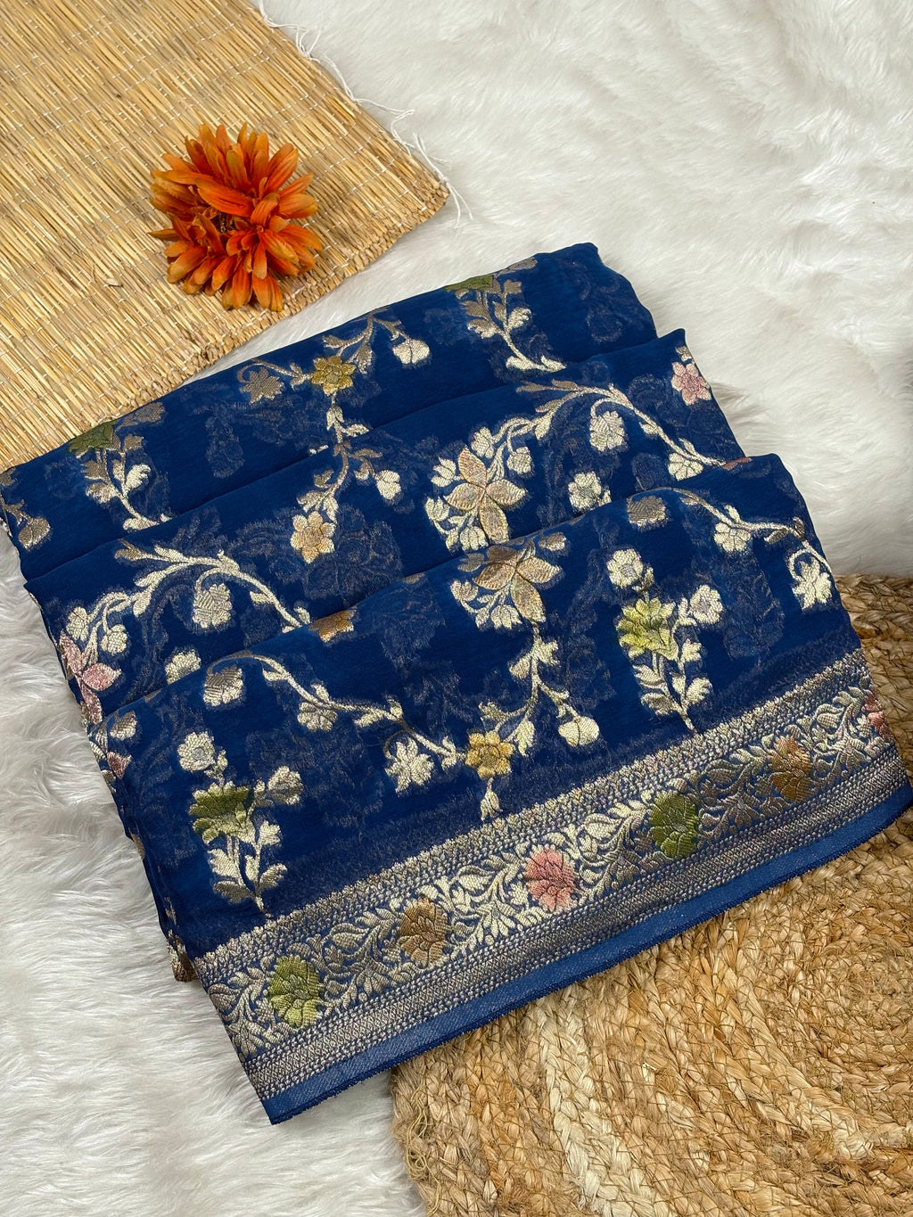 A folded navy blue saree with intricate floral weaving and a decorative border, styled on a white faux-fur surface with a woven mat and an orange flower prop.