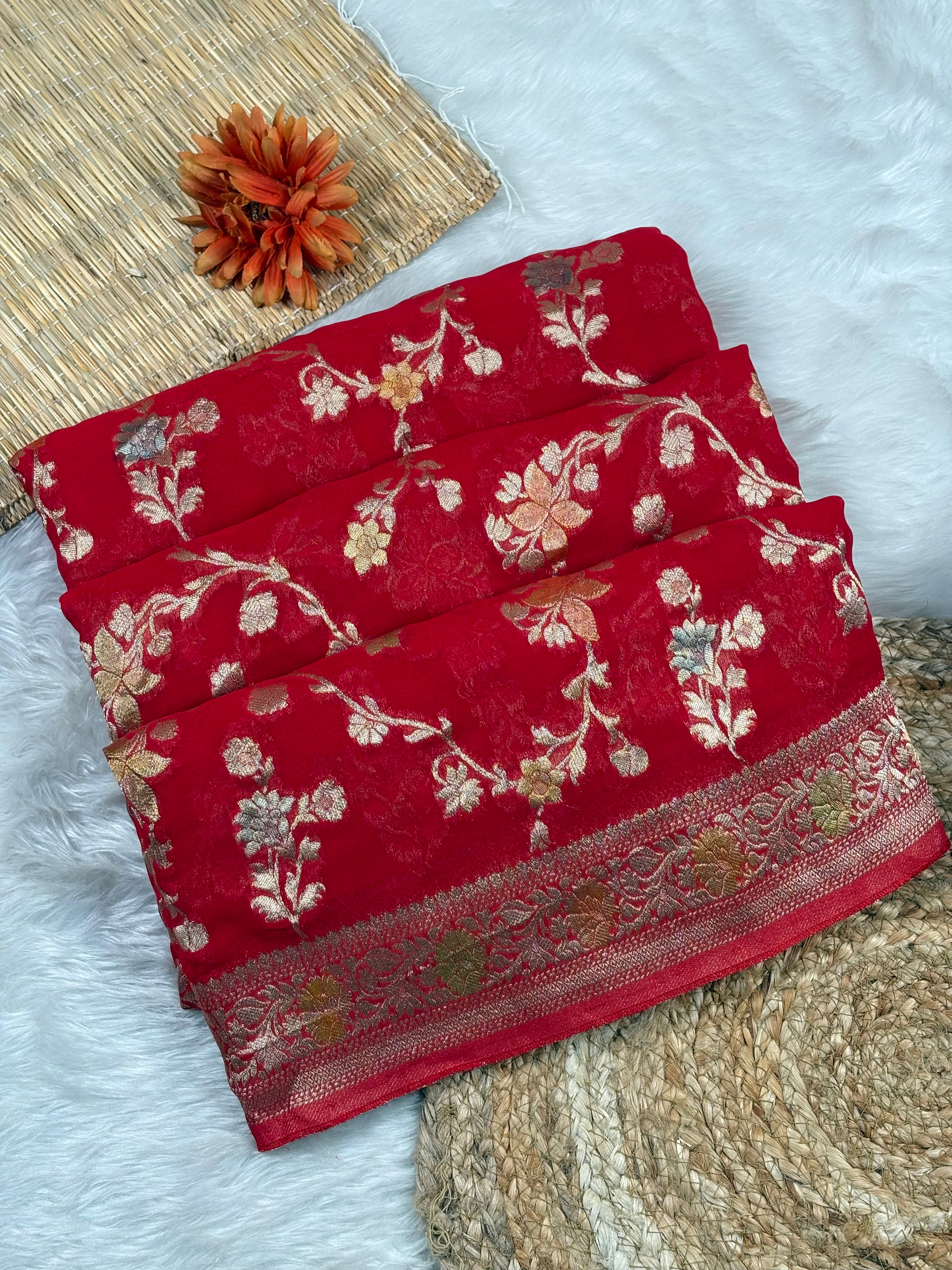 A folded red saree with intricate floral weaving and a decorative border, styled on a white faux-fur surface with woven mats and a dried orange flower.
