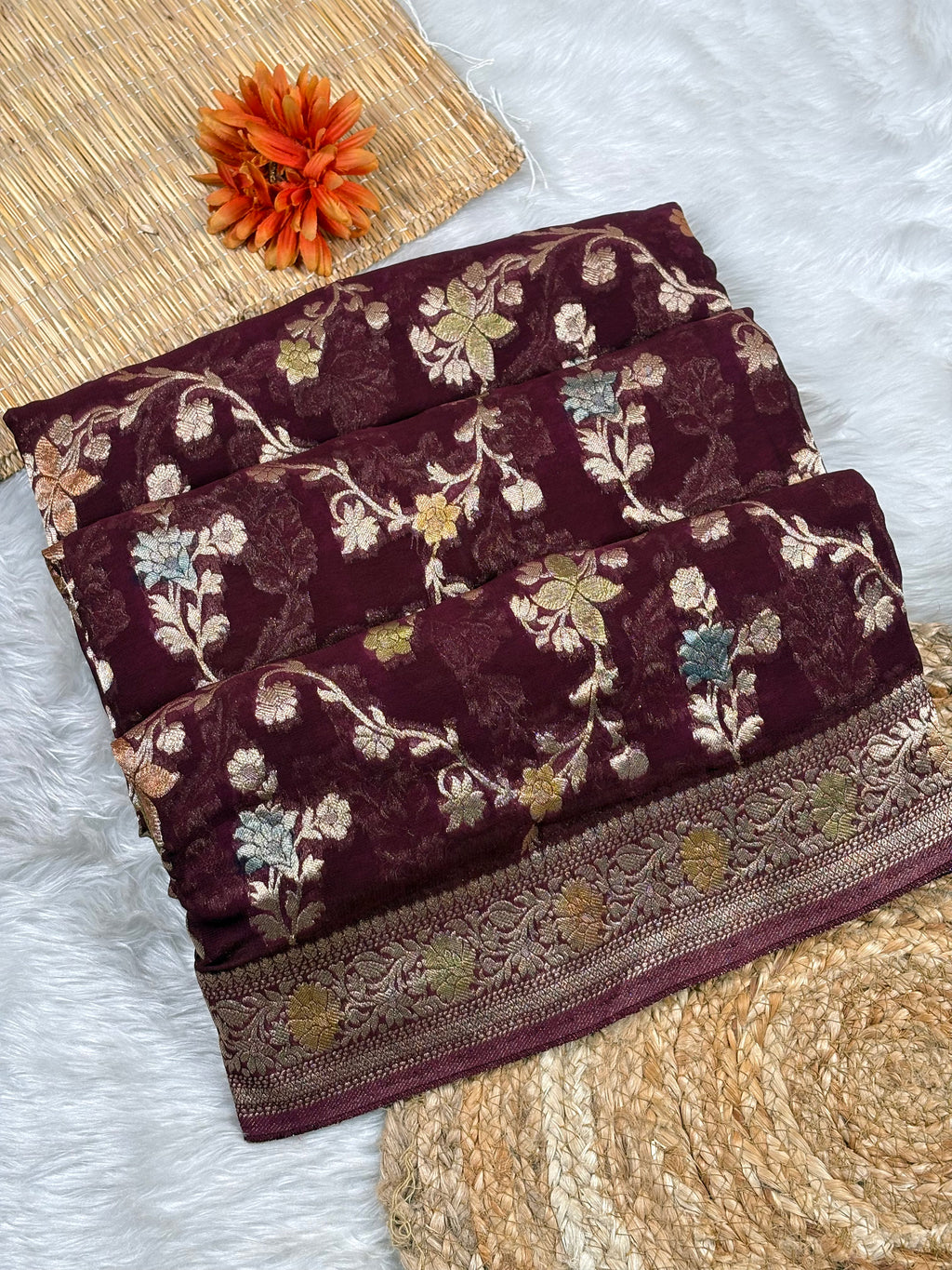 A folded deep maroon georgette saree with intricate floral and a decorative border, styled on a white faux fur surface with a woven mat and an orange flower prop.