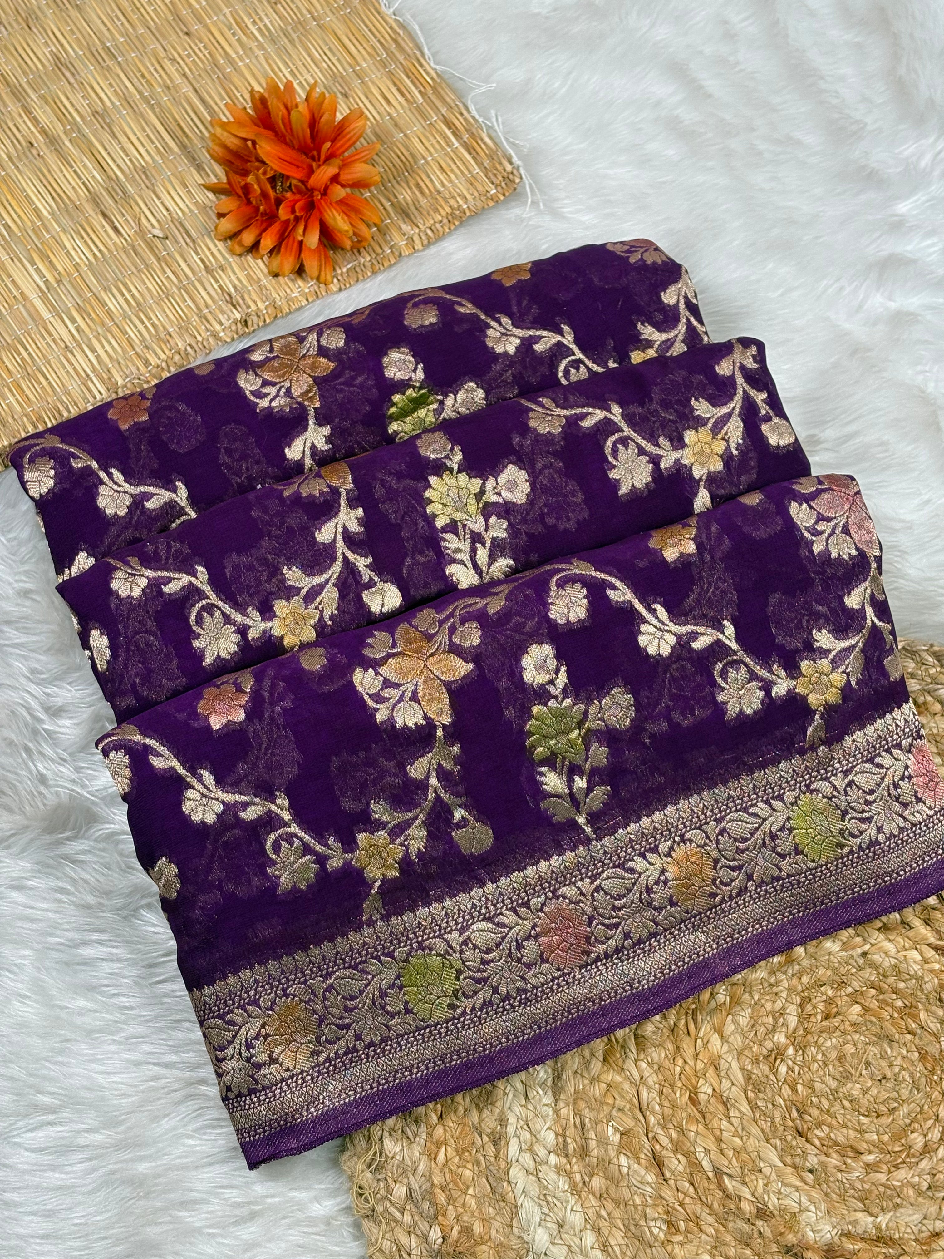 A folded dark purple Banarasi silk saree with intricate floral zari weaving and a wide decorative border, displayed on a white faux fur surface with a woven mat and orange flower prop.