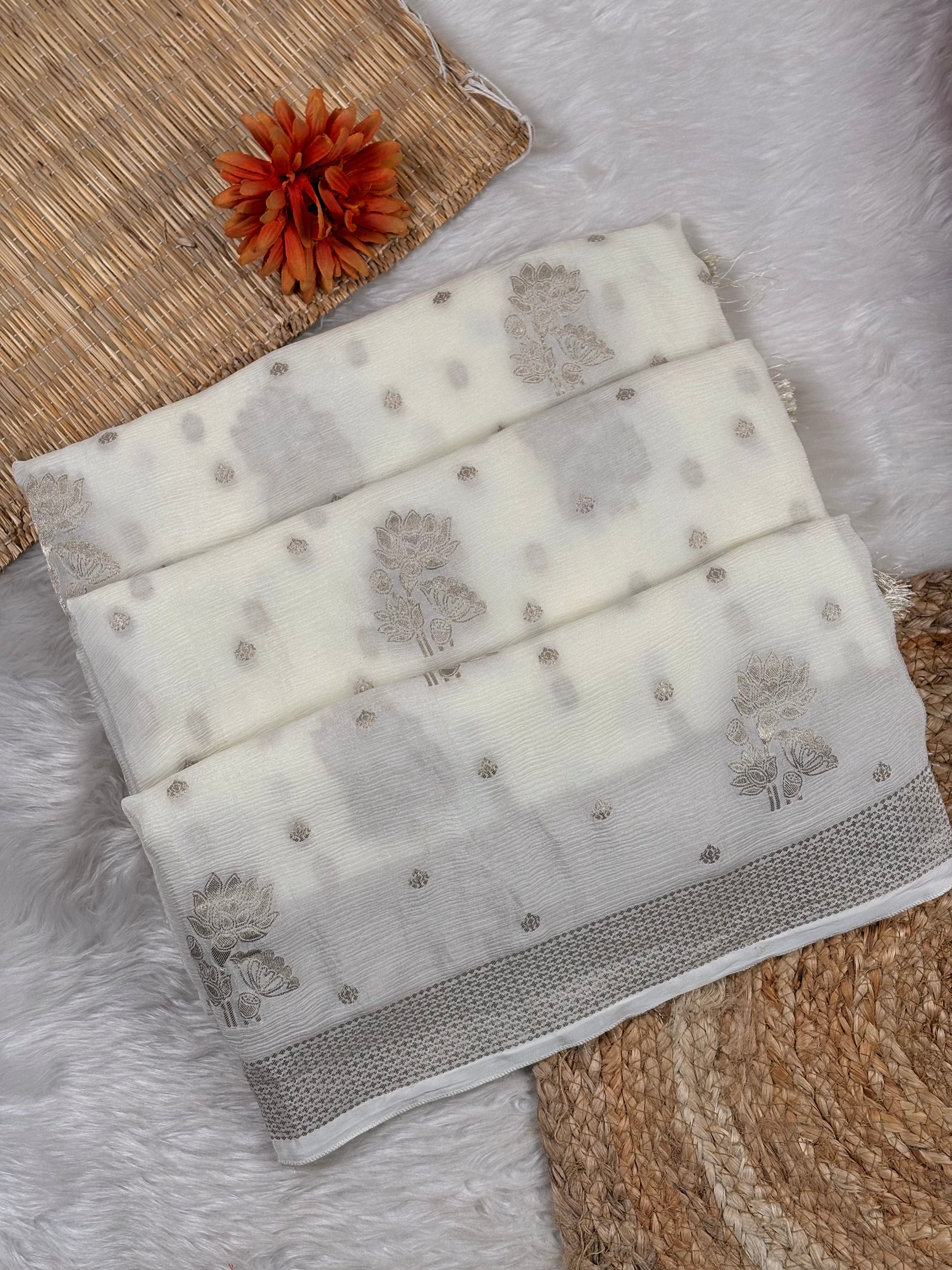 A folded off-white chinon saree with light gray floral and dot patterns, displayed on a white faux fur surface with a woven mat and a small orange flower as props.
