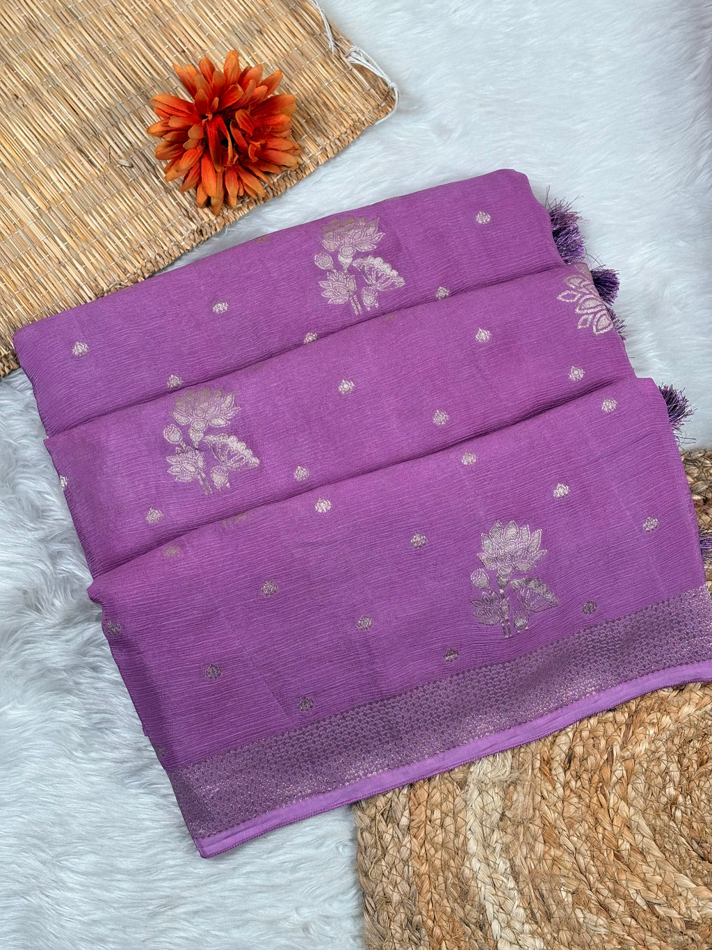A folded purple silk saree with water gold floral and dot motifs, displayed on a white faux fur surface with a woven mat and orange flower prop.