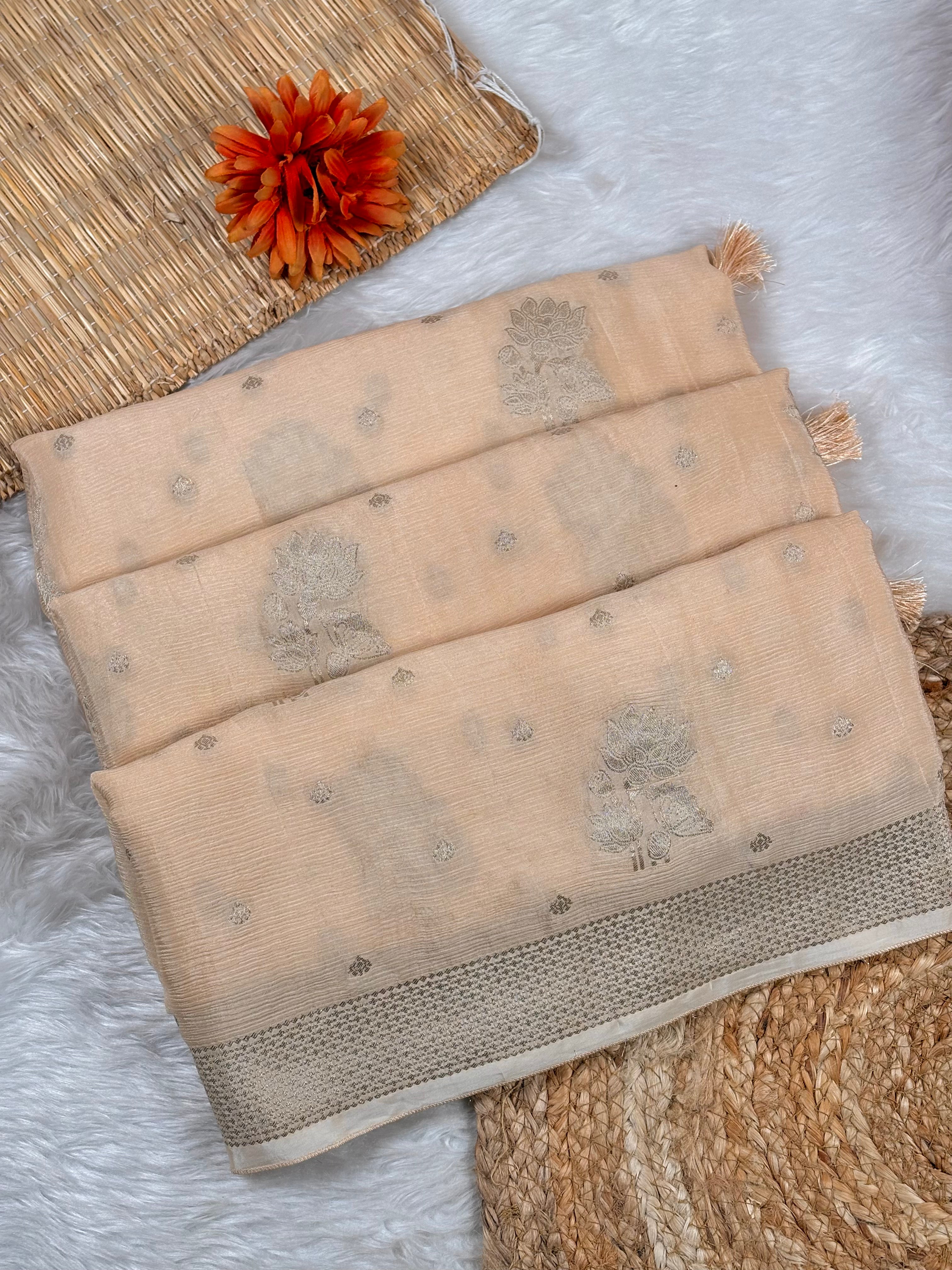 A folded beige saree with subtle floral motifs and a contrasting border, styled on a white faux fur surface with a woven mat and an orange flower accent.