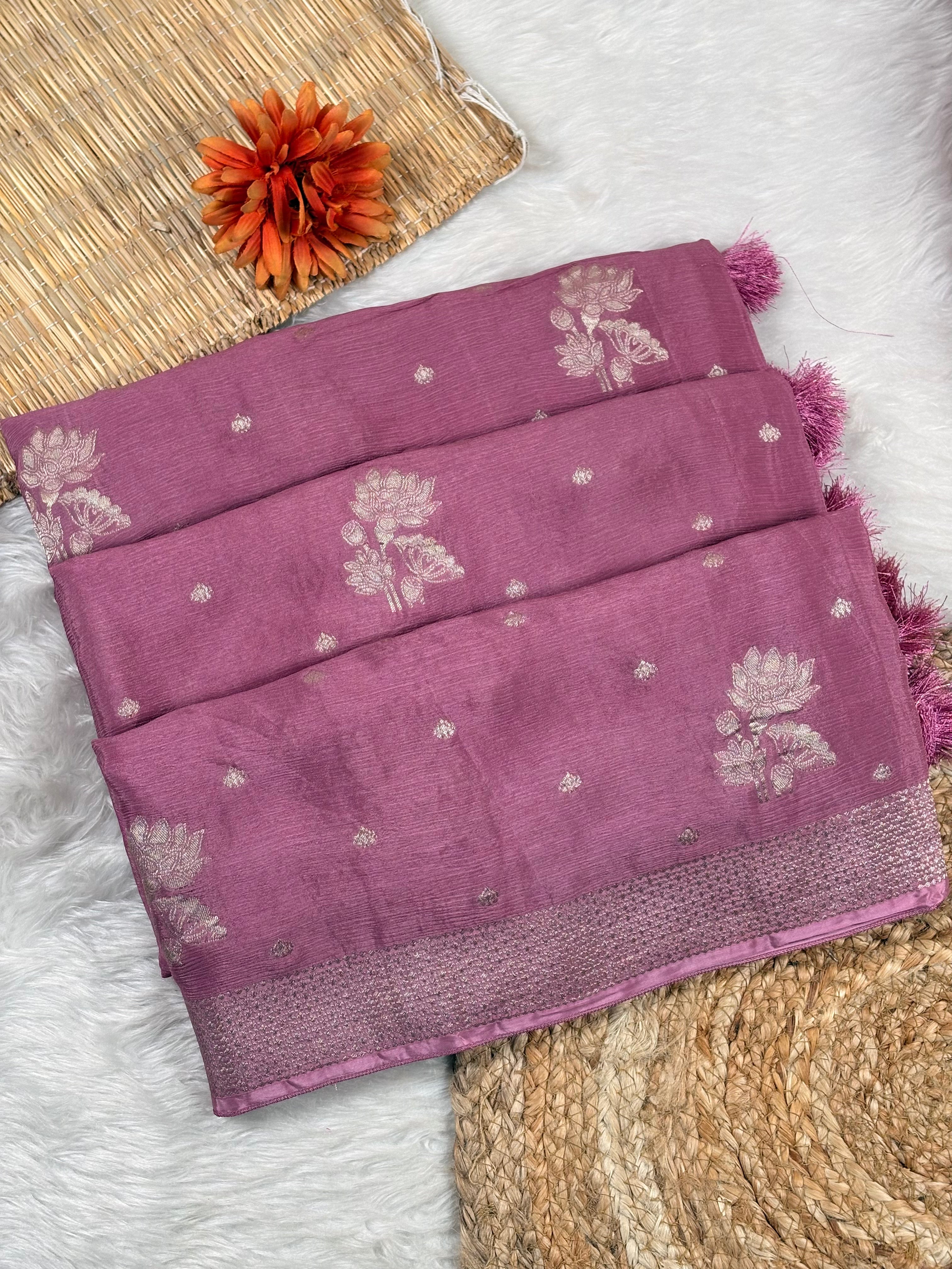 A folded dusty purple silk saree with white floral and dot motifs, displayed on a white faux fur rug alongside a woven mat and an orange flower.