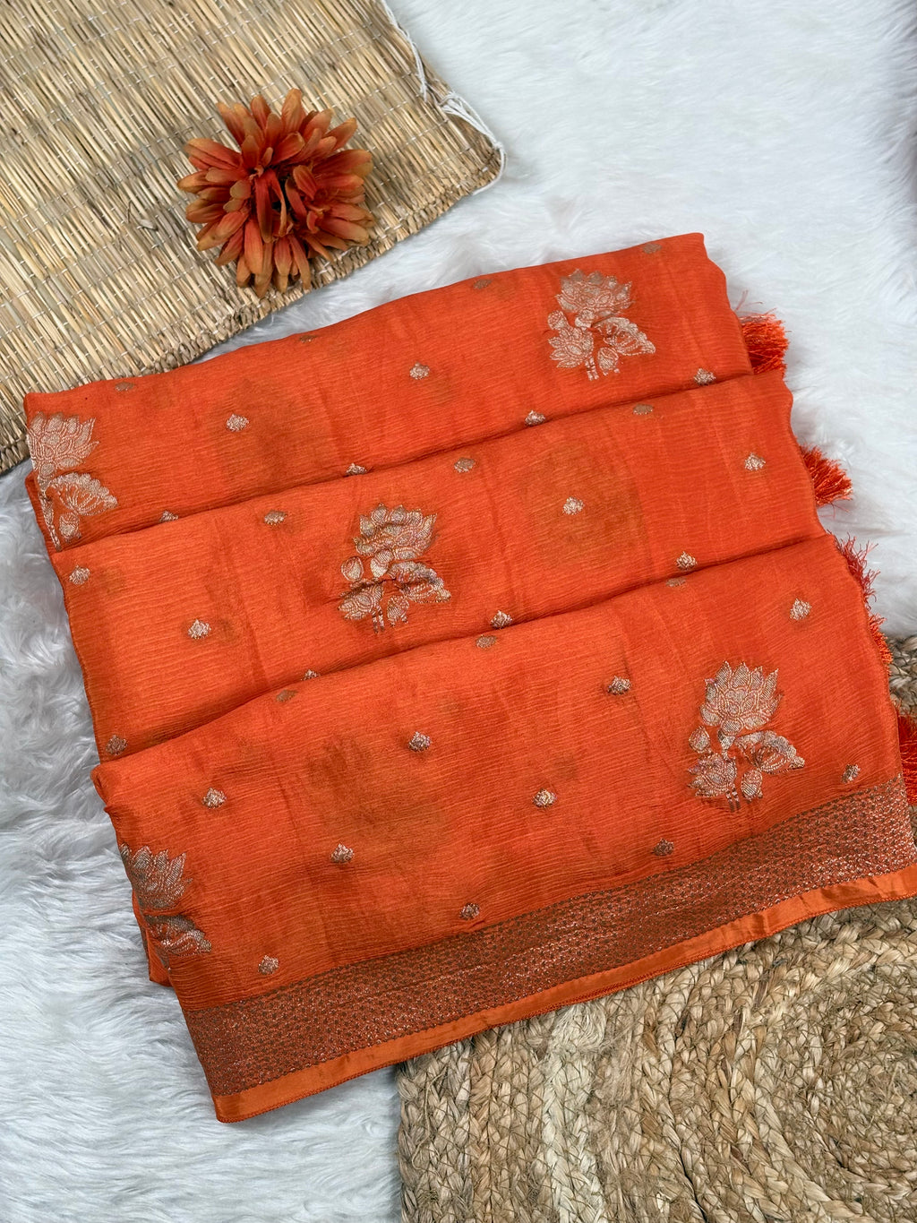An orange saree with water gold floral motifs and small metallic water gold dots, laid flat on a white faux fur surface with decorative woven mats and a dried flower.