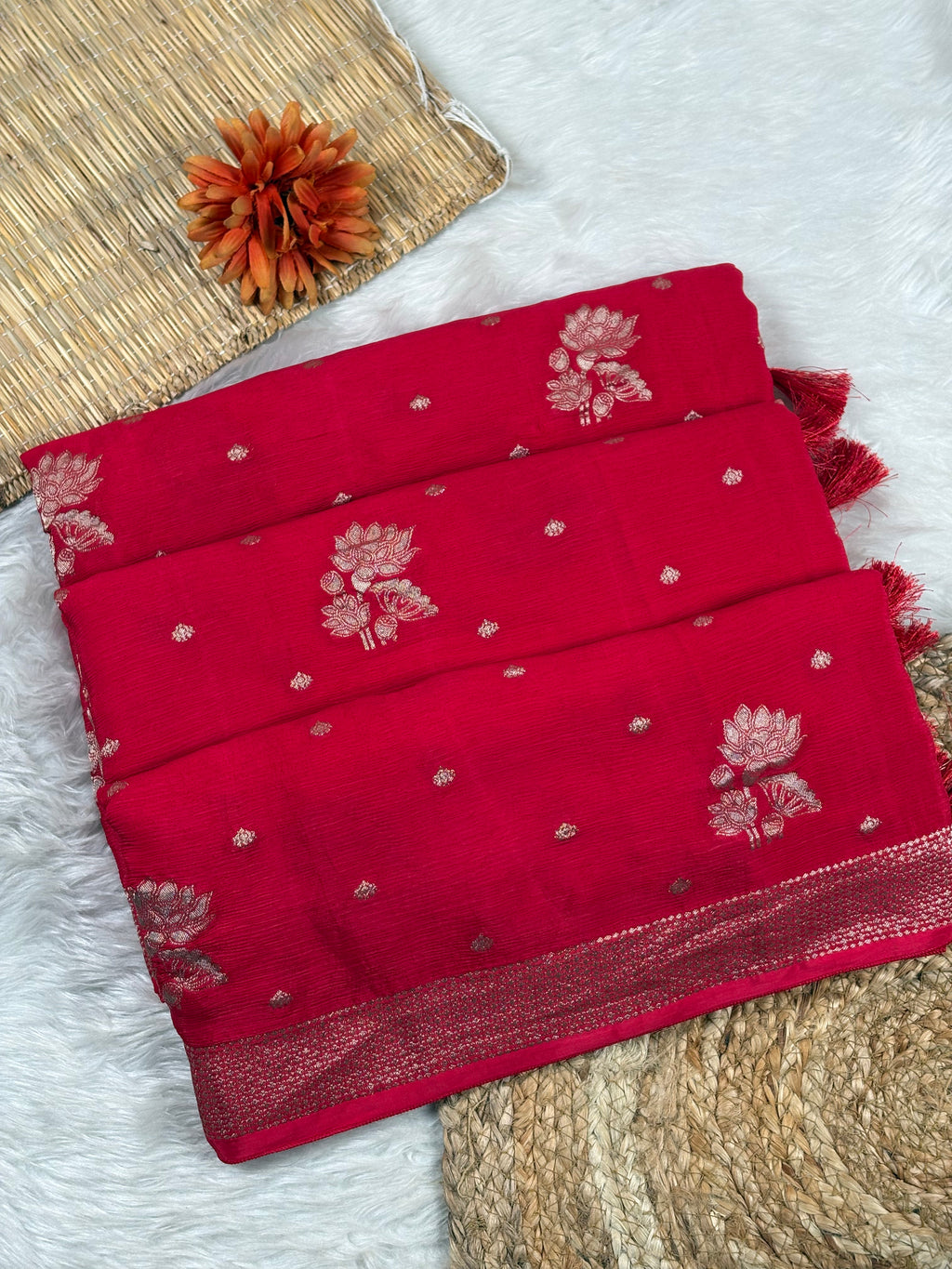 A folded red saree with water gold floral motifs and a water gold border, displayed on a white faux-fur surface with woven props. 