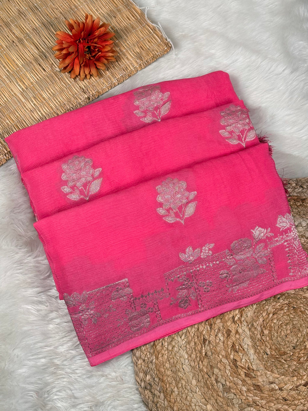 A folded bright pink saree with light pink floral motifs and a decorative border, styled on a white faux-fur surface with woven mat props and a small orange flower.